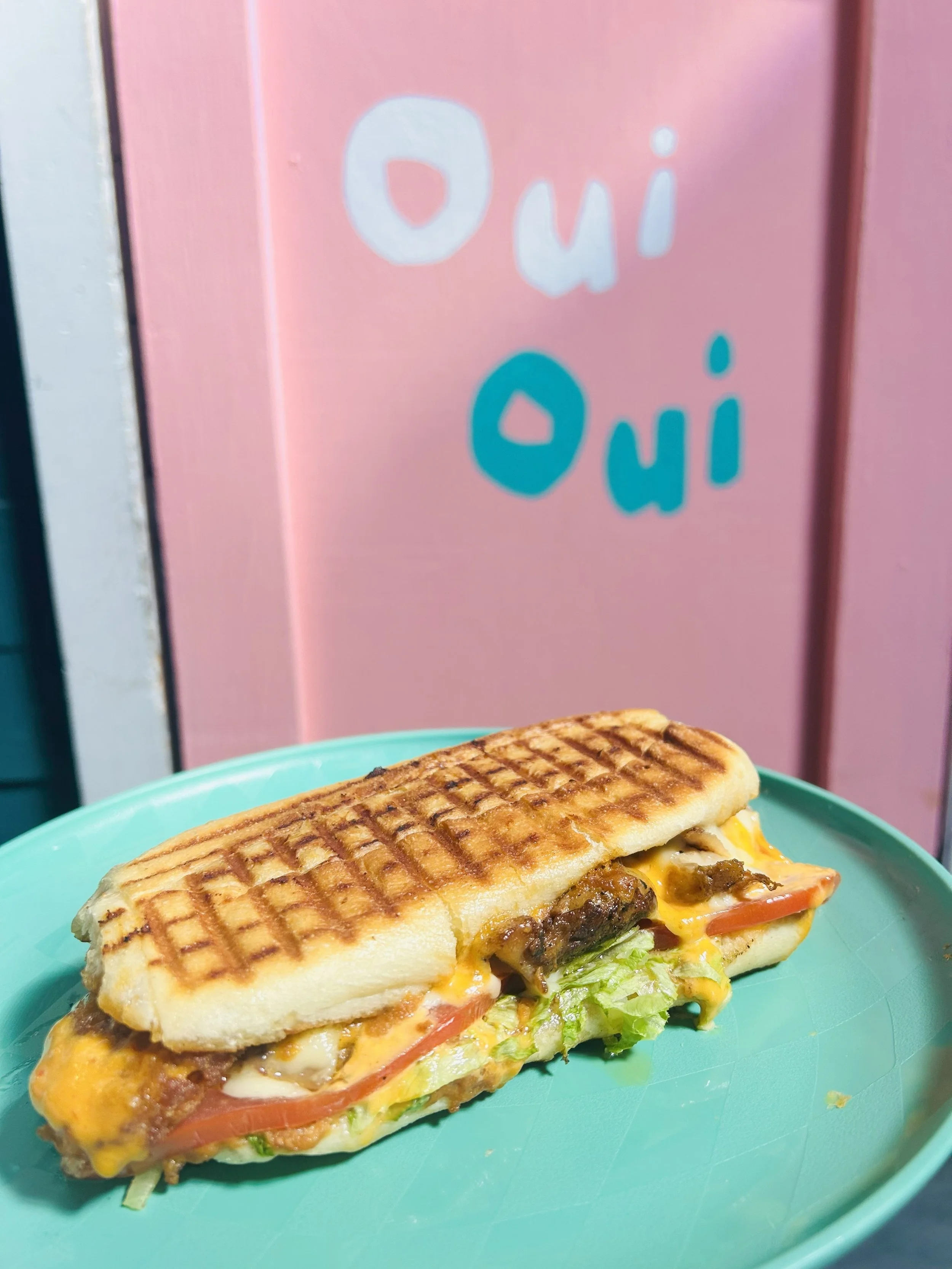 A grilled sandwich with cheese, lettuce, tomato, and beef on a blue plate in front of a pink background with the words "Oui" and "Oui" written on it.