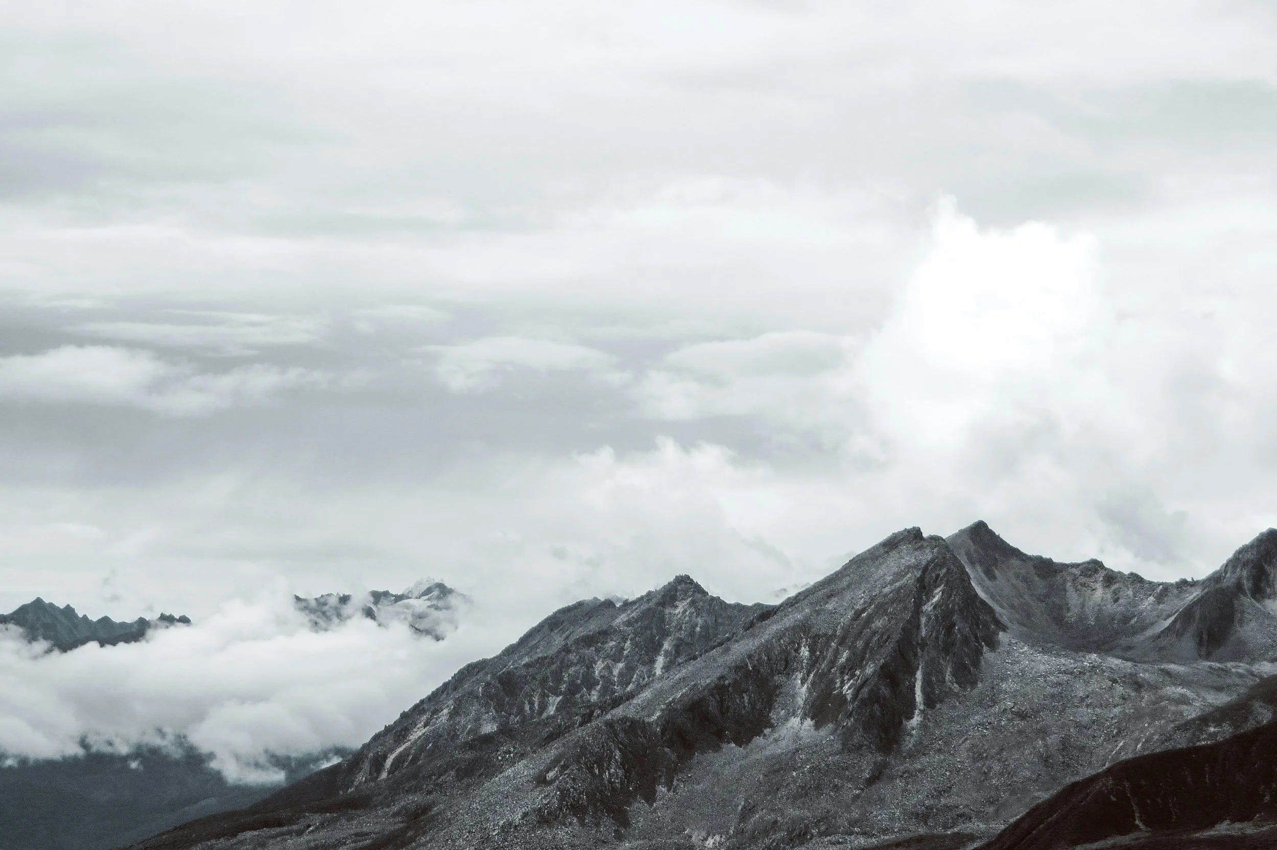 Mountains with rocky terrain in the foreground, clouds and mist surrounding the peaks, and an overcast sky.