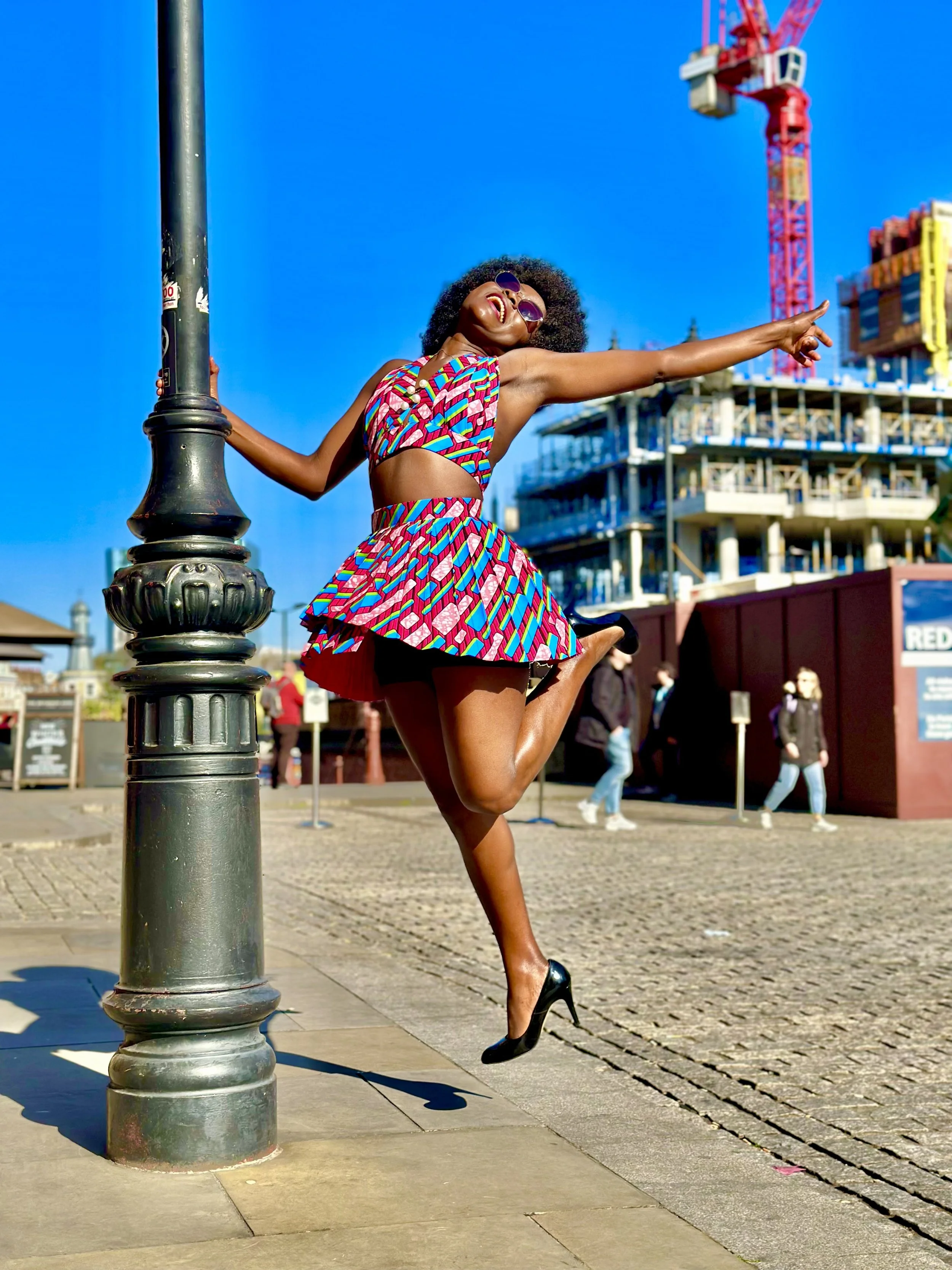 Person in colorful outfit posing energetically by a lamppost, with a construction crane and building in the background.