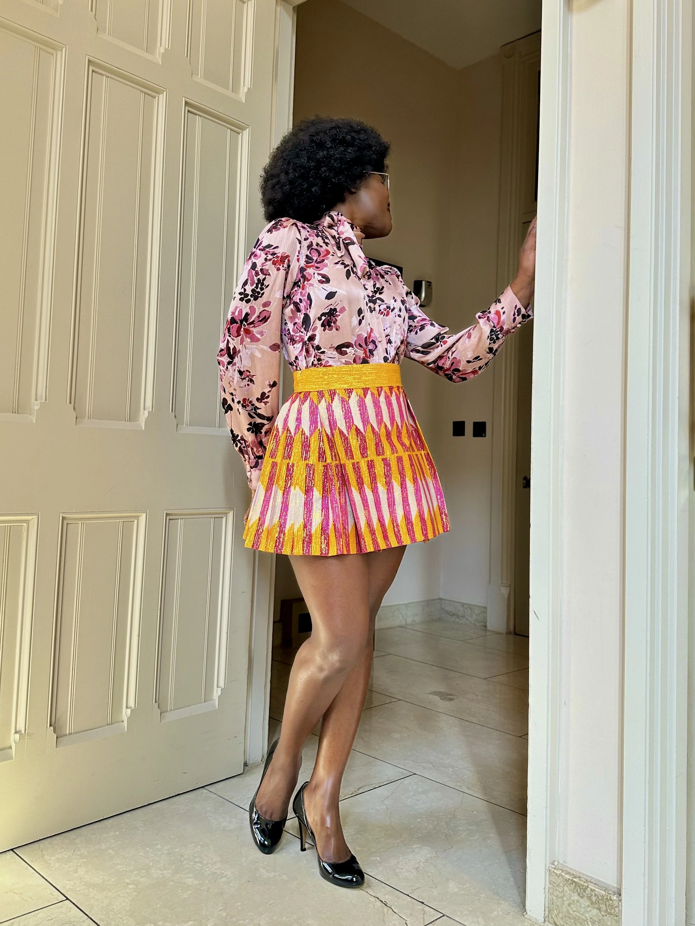 Person wearing a floral blouse and colorful striped skirt, standing in a doorway with high heels.