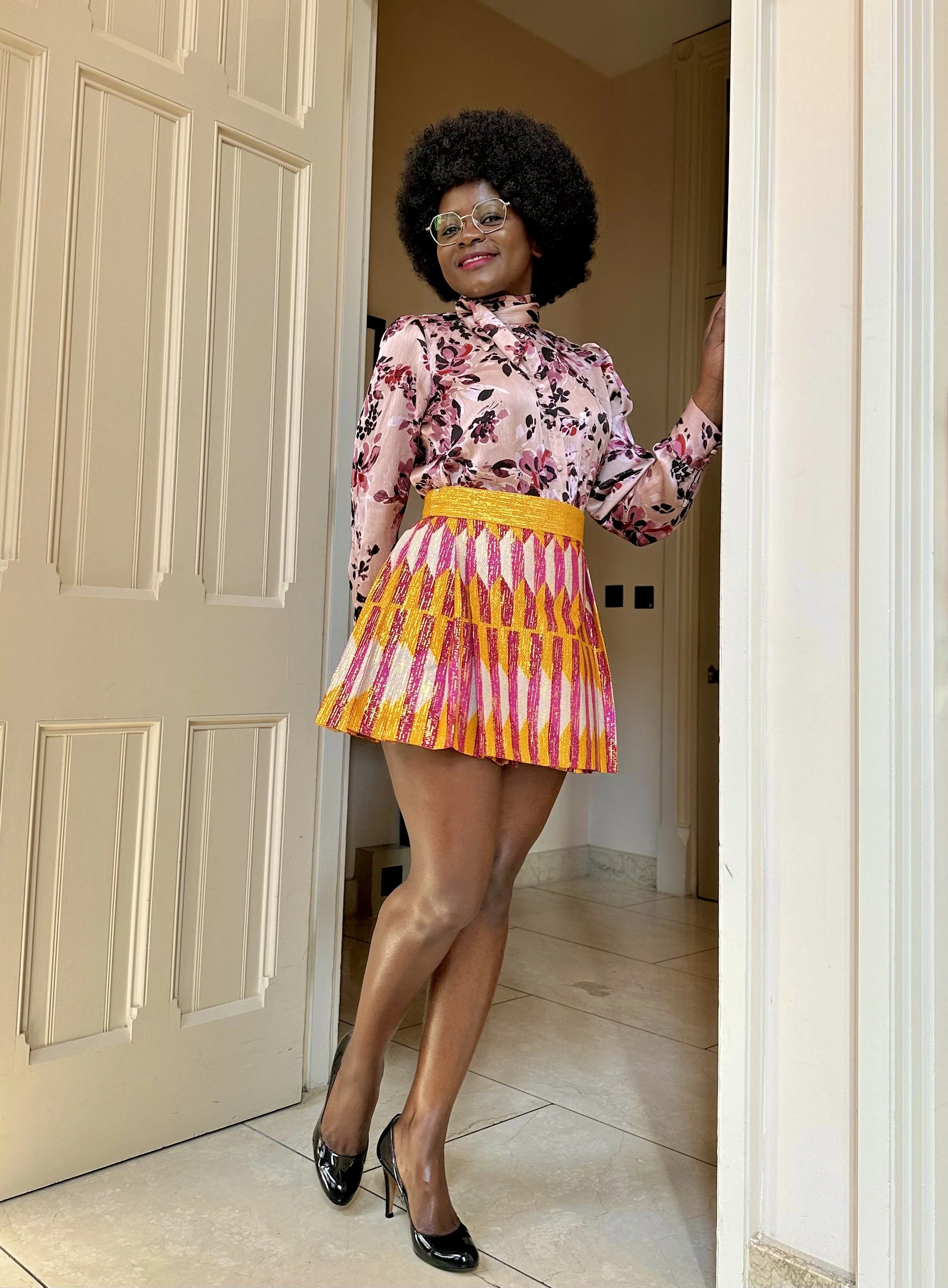 Person wearing a floral blouse, colorful pleated skirt, and black high heels, standing in a doorway.