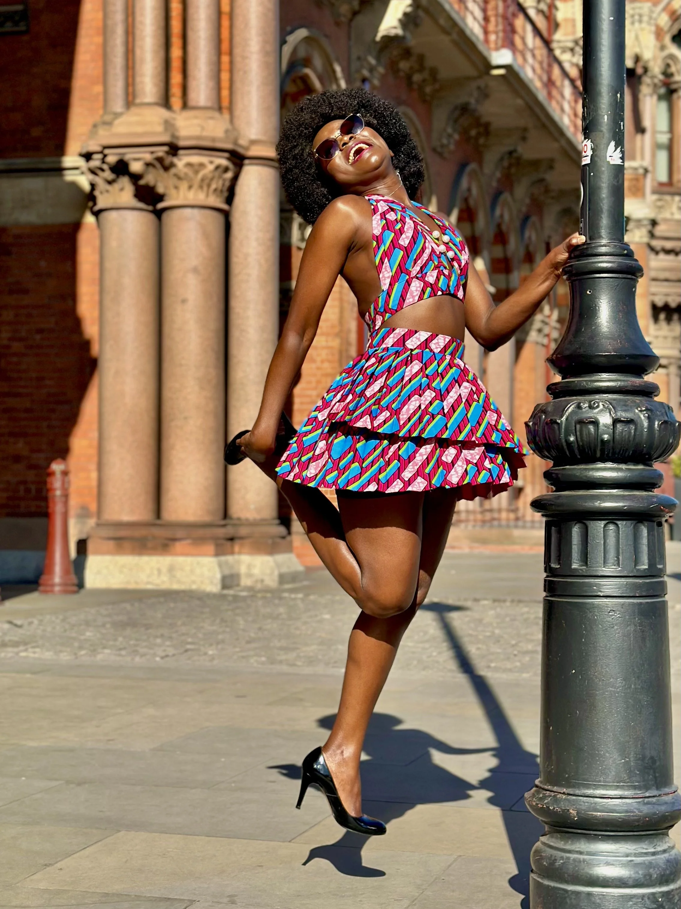 Person in colorful dress joyfully jumping next to a lamp post, urban architectural background.
