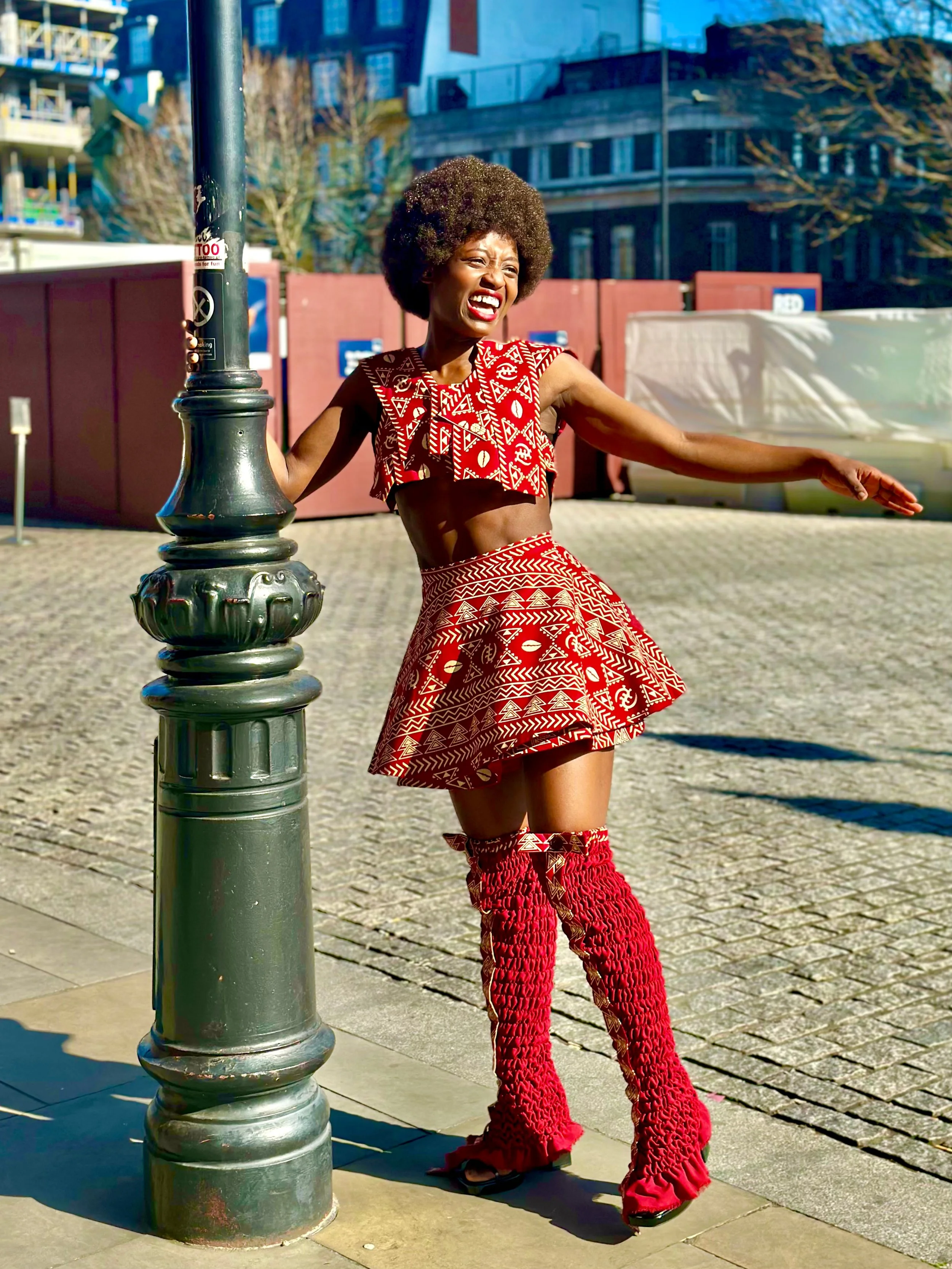 Person wearing red patterned outfit with afro hairstyle, posing by a lamppost on a sunny urban street.