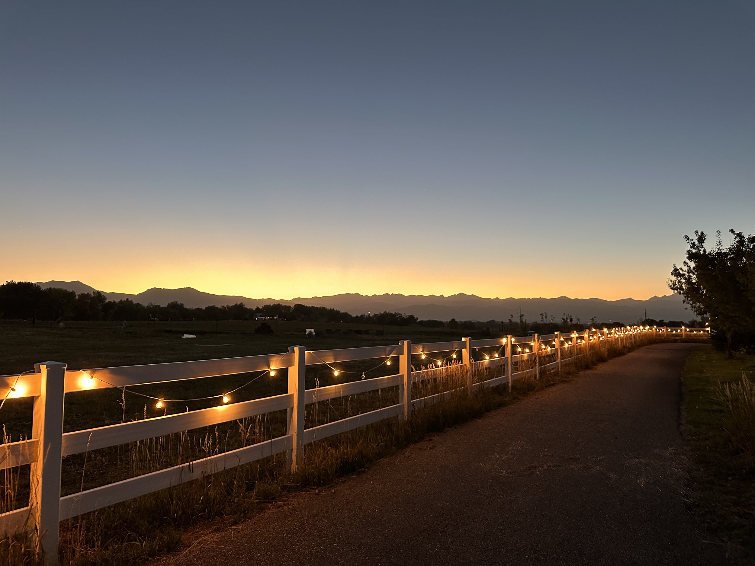 A sunset scene with a path running alongside a white fence decorated with string lights, with mountains in the background and a clear sky transitioning from light to dark.