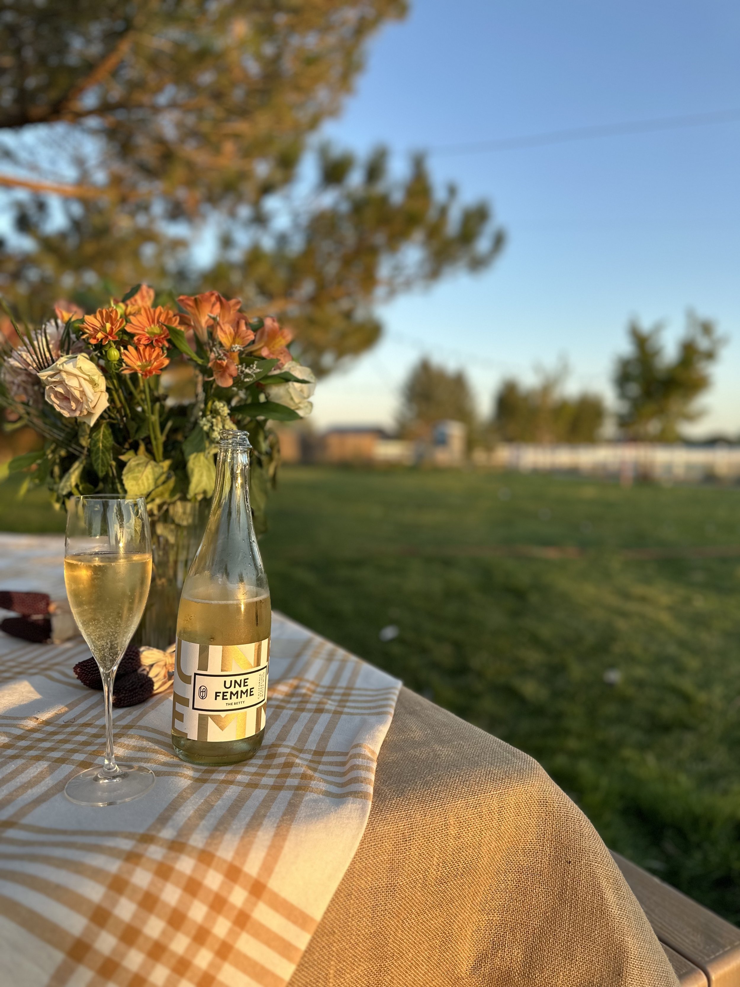 A table set outdoors with a bouquet of flowers, a glass of sparkling wine, and a bottle of wine labeled 'Une Femme' on a checkered tablecloth, with a grassy lawn and trees in the background during sunset.