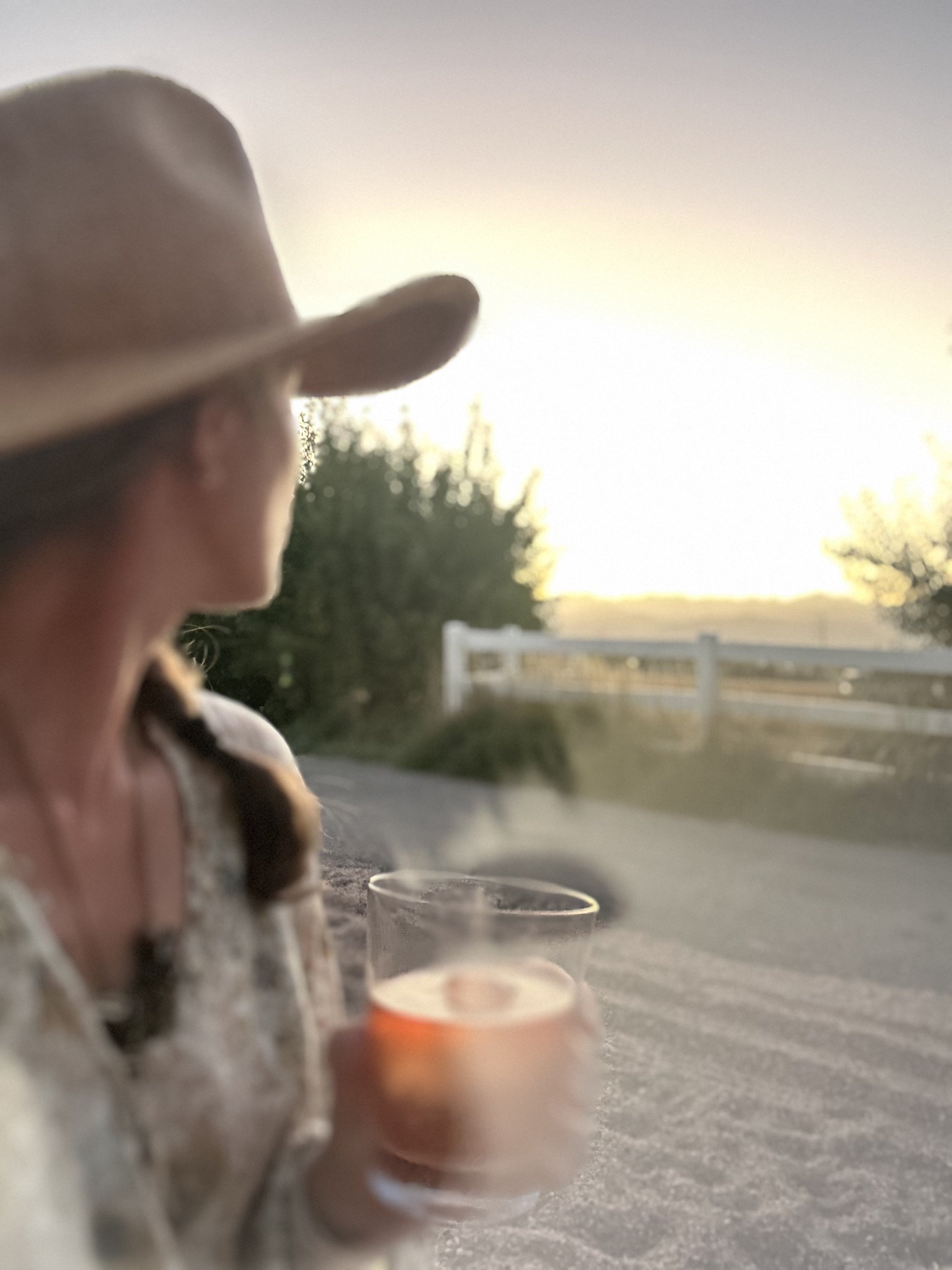 A woman wearing a cowboy hat holding a glass of wine or champagne or cocktail in a scenic outdoor setting at sunset.