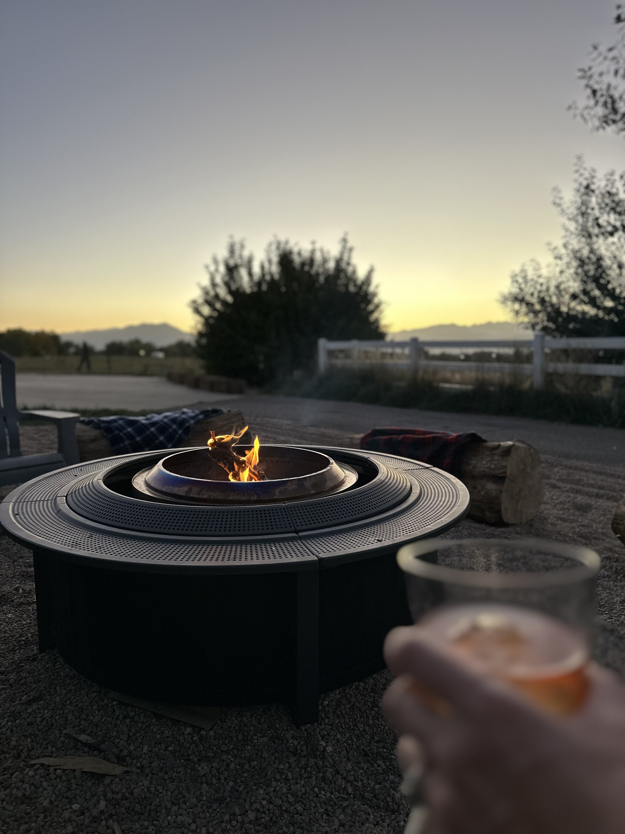 Person holding a drink near a fire pit outdoors at sunset with benches and trees in the background.