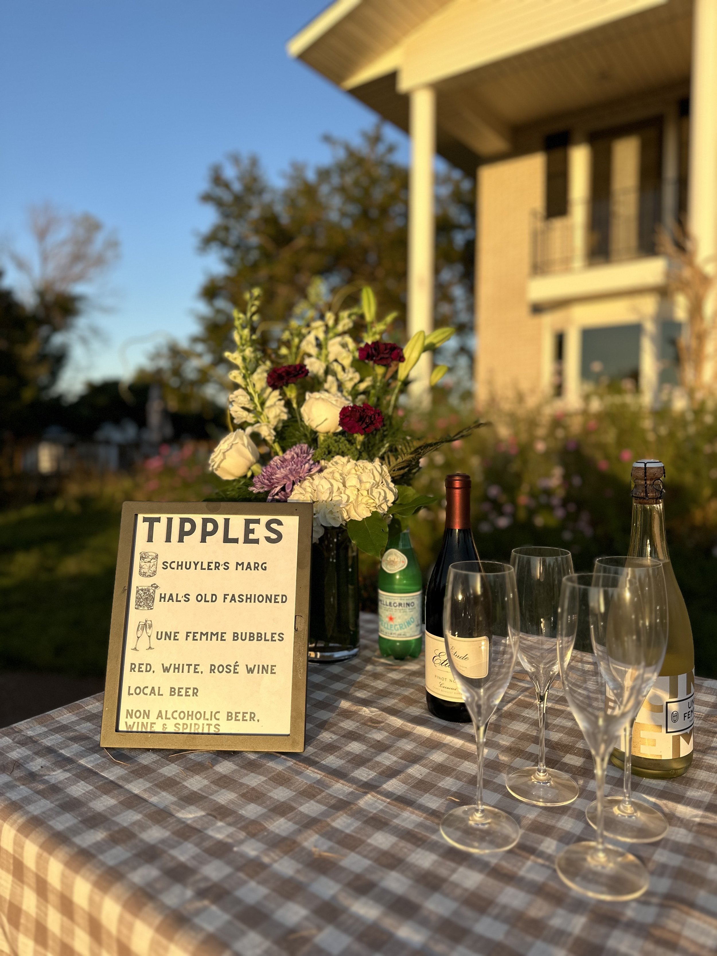 A table set for an outdoor gathering with a floral centerpiece, bottles of wine and non-alcoholic beverages, four empty champagne glasses, and a menu card titled "Tipples" listing various drinks, on a checkered tablecloth with a house and trees in th