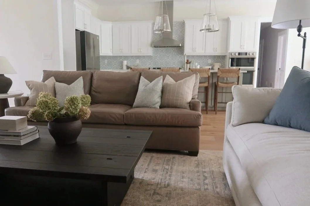 Living room with beige and white sofas, a black coffee table with a flower arrangement, and an open kitchen with white cabinets and a patterned backsplash in the background.