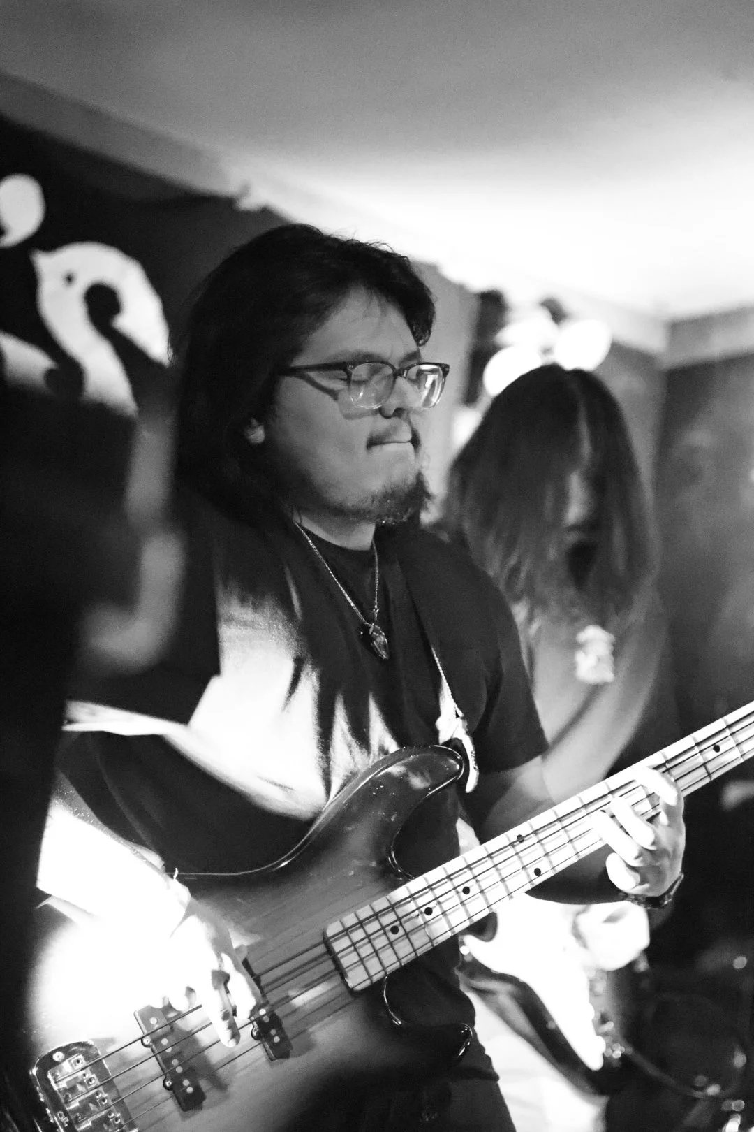 A man with glasses and long hair playing an electric bass guitar in a dimly lit venue.