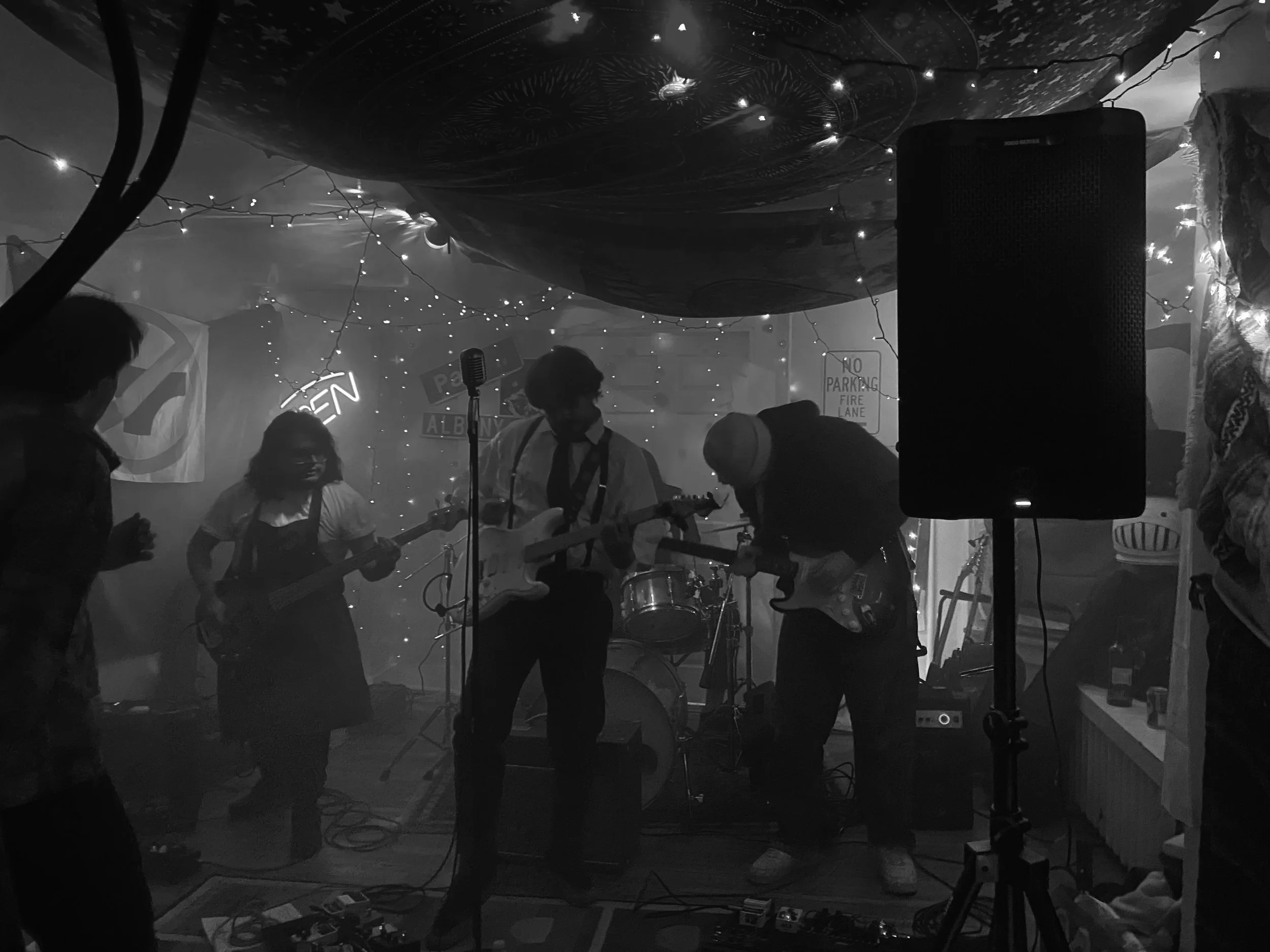 A black-and-white photo of a band performing in a dimly lit venue with string lights hanging from the ceiling. The band members are playing guitars and drums, with a microphone visible in front. Audience members are seen in the foreground, and a larg