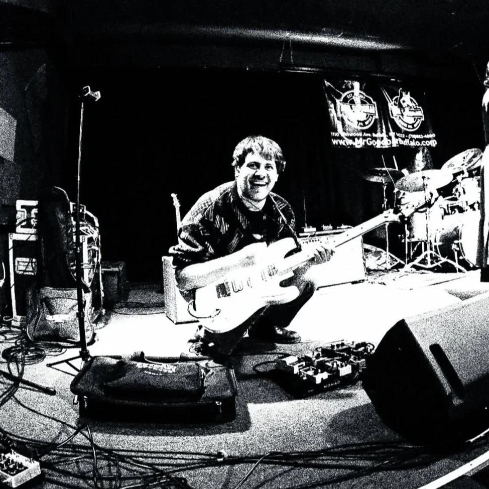 A man kneeling on stage with an electric guitar, smiling, with a drum set and banners in the background, surrounded by musical equipment.