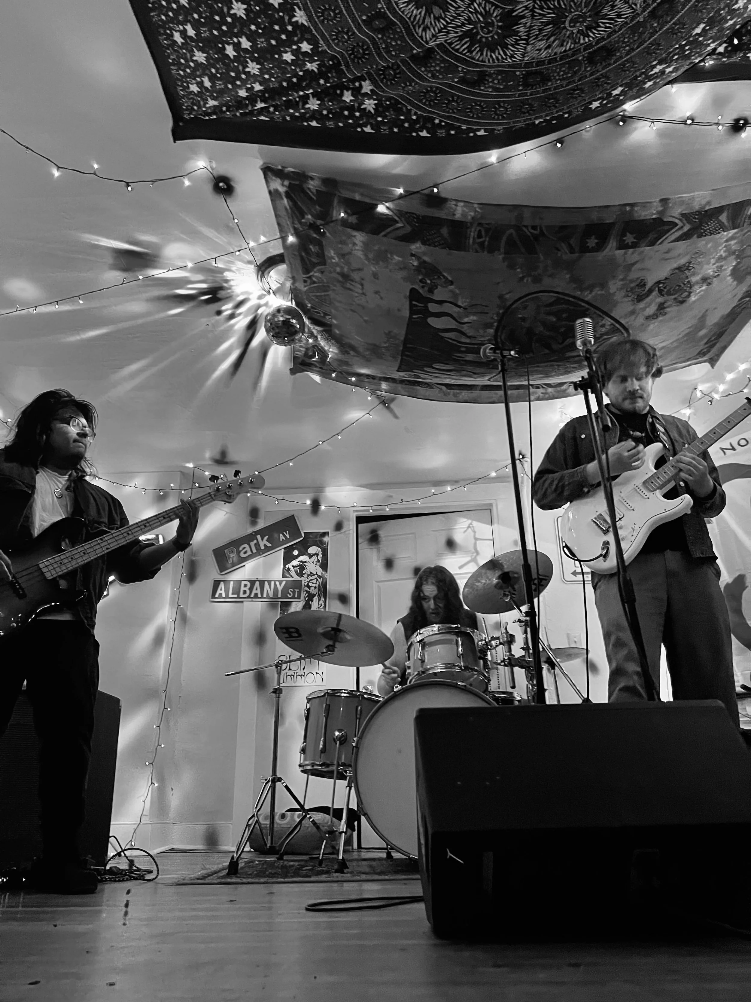 A black and white photo of a three-member band performing on a small stage. Two guitarists and a drummer are visible. The stage is decorated with string lights and patterned fabric on the ceiling and walls. There are street signs reading 'Park AV' an
