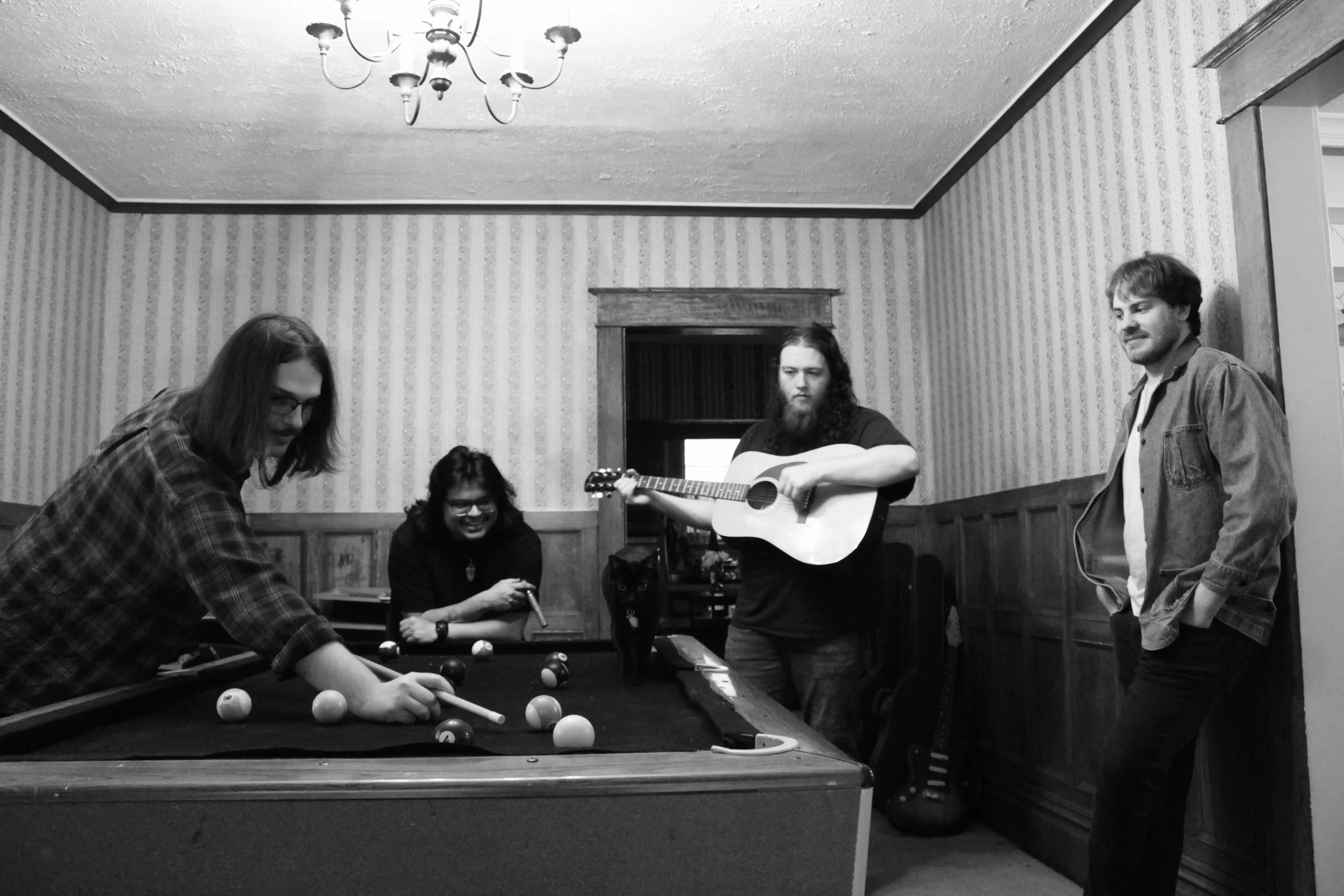 A group of five people are gathered around a pool table in a room with striped wallpaper. One person is taking a shot, another is playing guitar, and the others are observing and socializing.