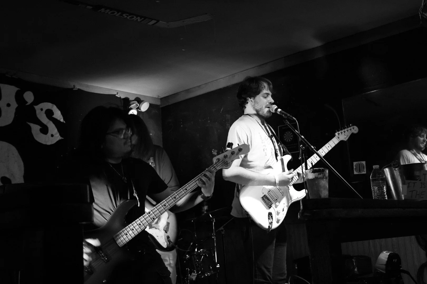 Black and white photo of a band performing on stage, with a male guitarist and vocalist in the foreground, women playing bass and keyboard in the background, on a dimly lit stage in a small venue.