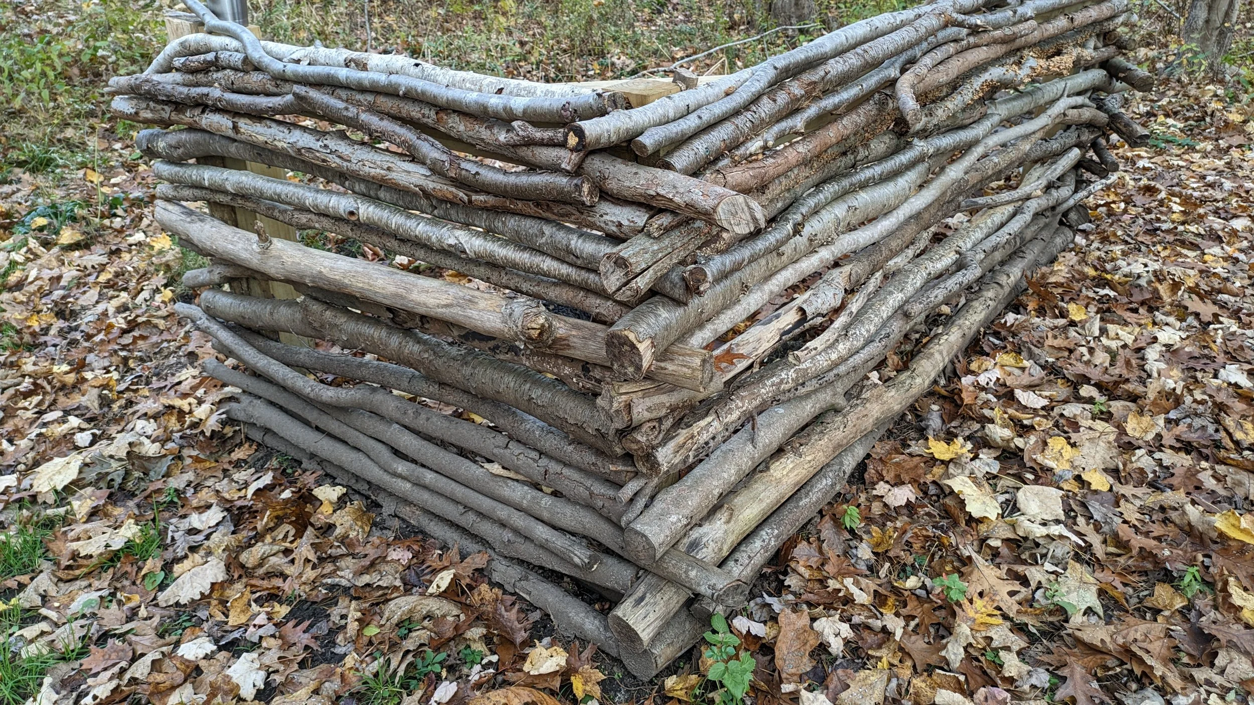 Wooden sticks stacked in a rectangular shape on a forest floor covered with fallen leaves.