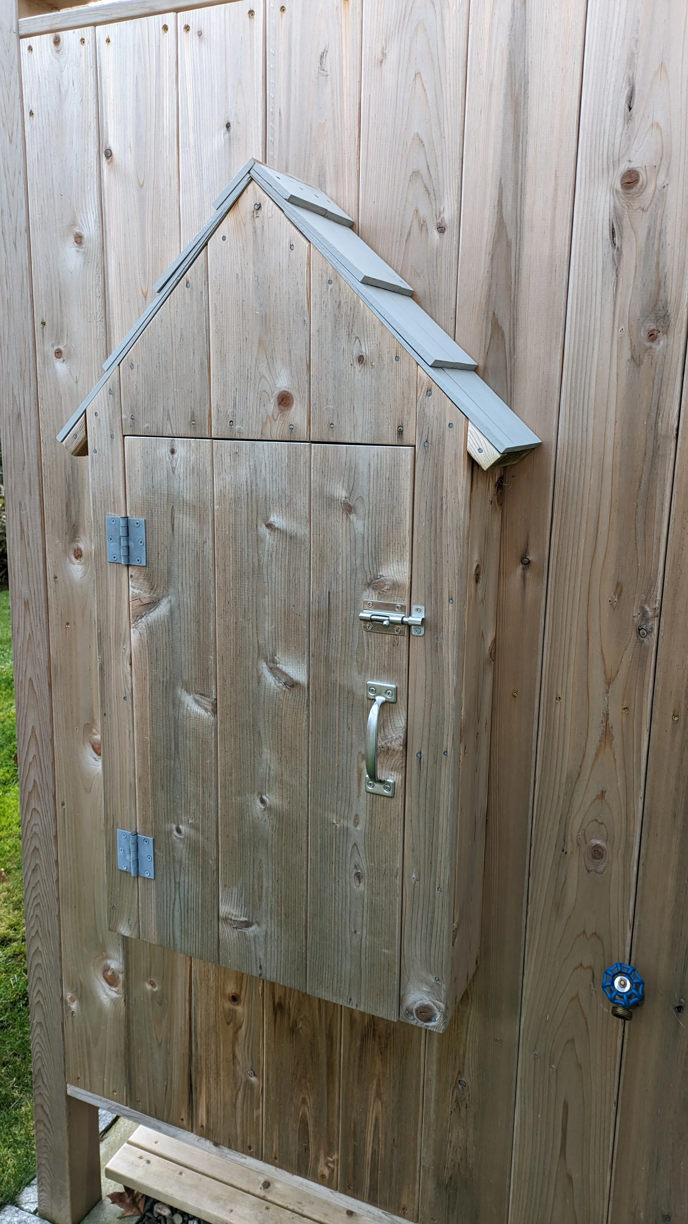 A small wooden door with a handle, hinges, and a latch, installed on a wooded outdoor wall, with a small roof above the door.