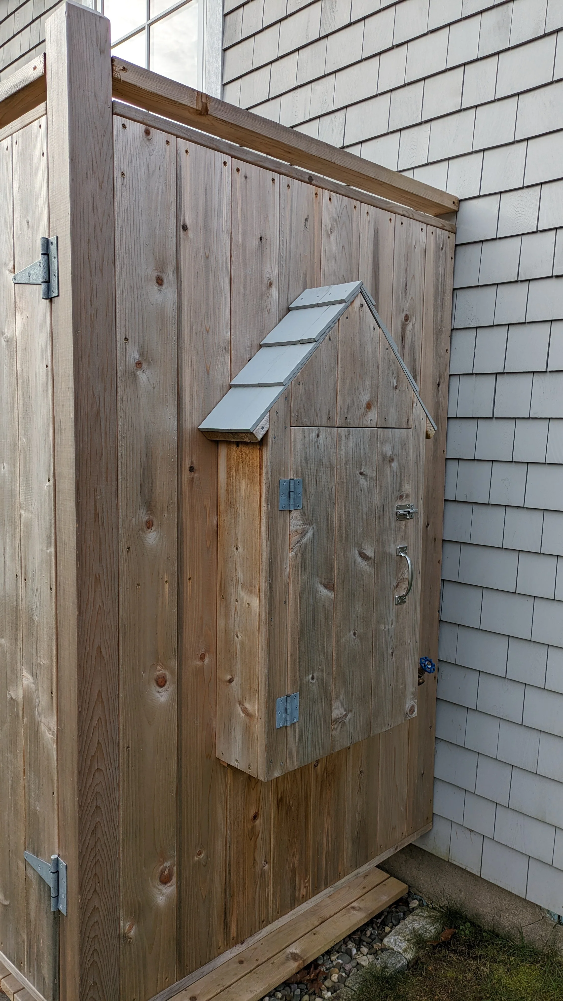 Wooden outdoor structure with a door and a small roof, attached to a house with gray siding.