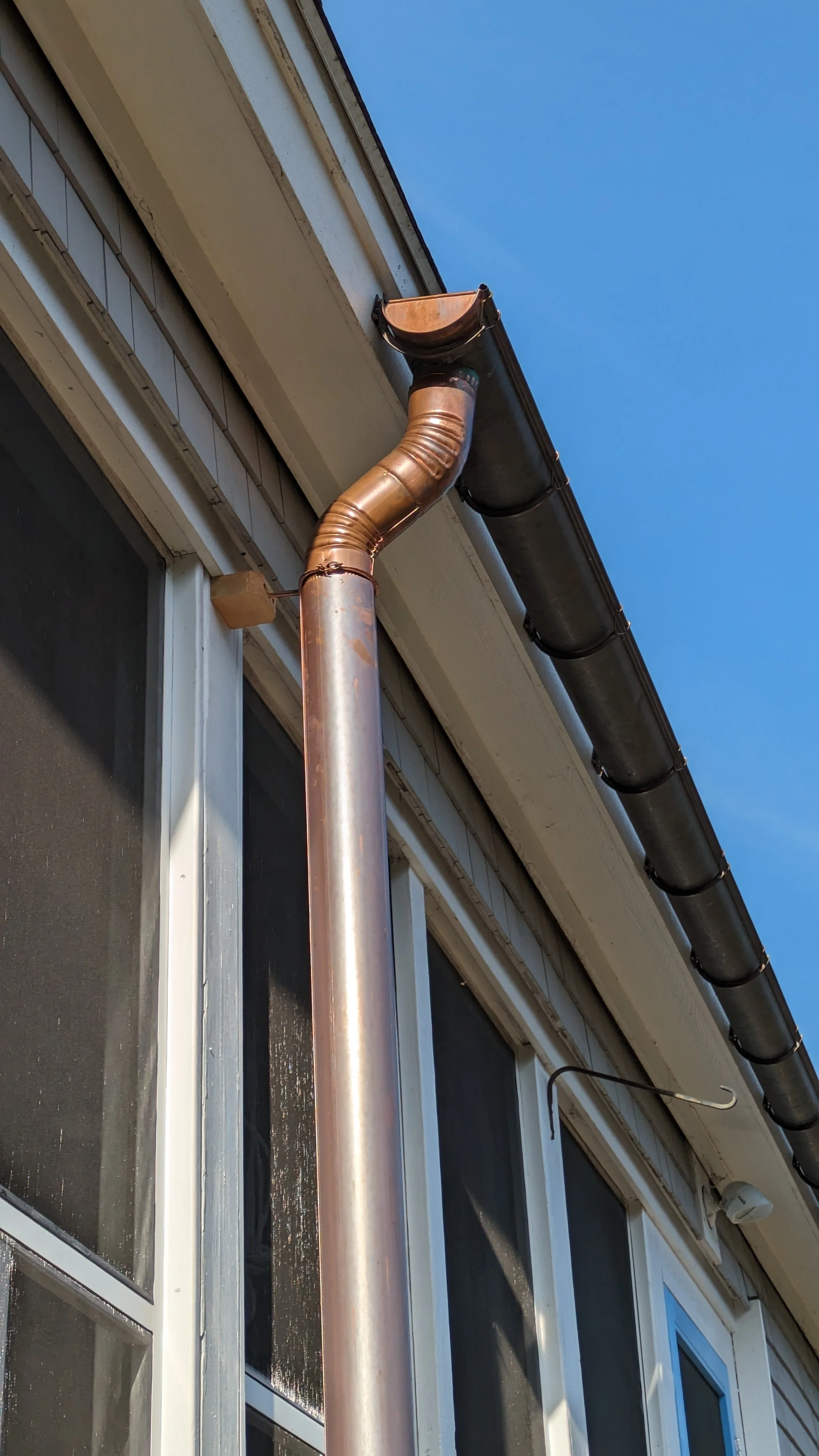 Close-up of a house's exterior showing a copper-colored downspout connected to a black gutter system, with a window and siding visible beneath a clear blue sky.