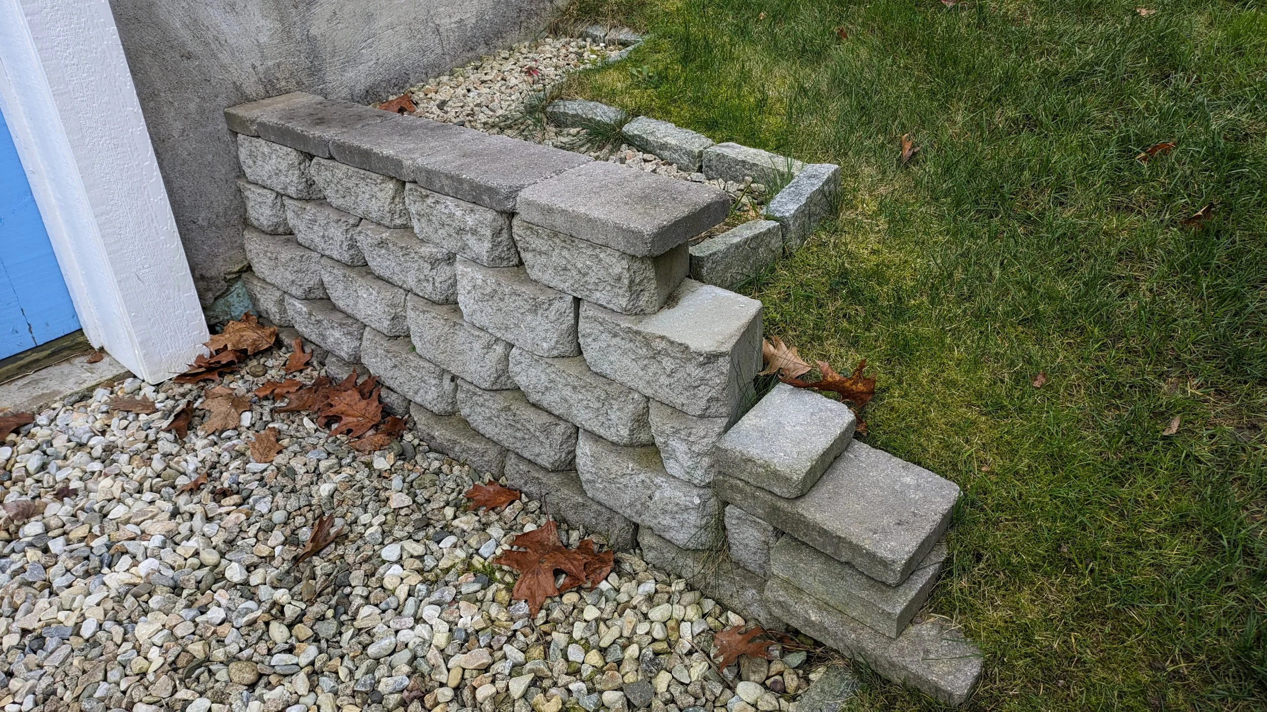 Stacked stone bricks forming a small wall near a building with a grass lawn and fallen autumn leaves.