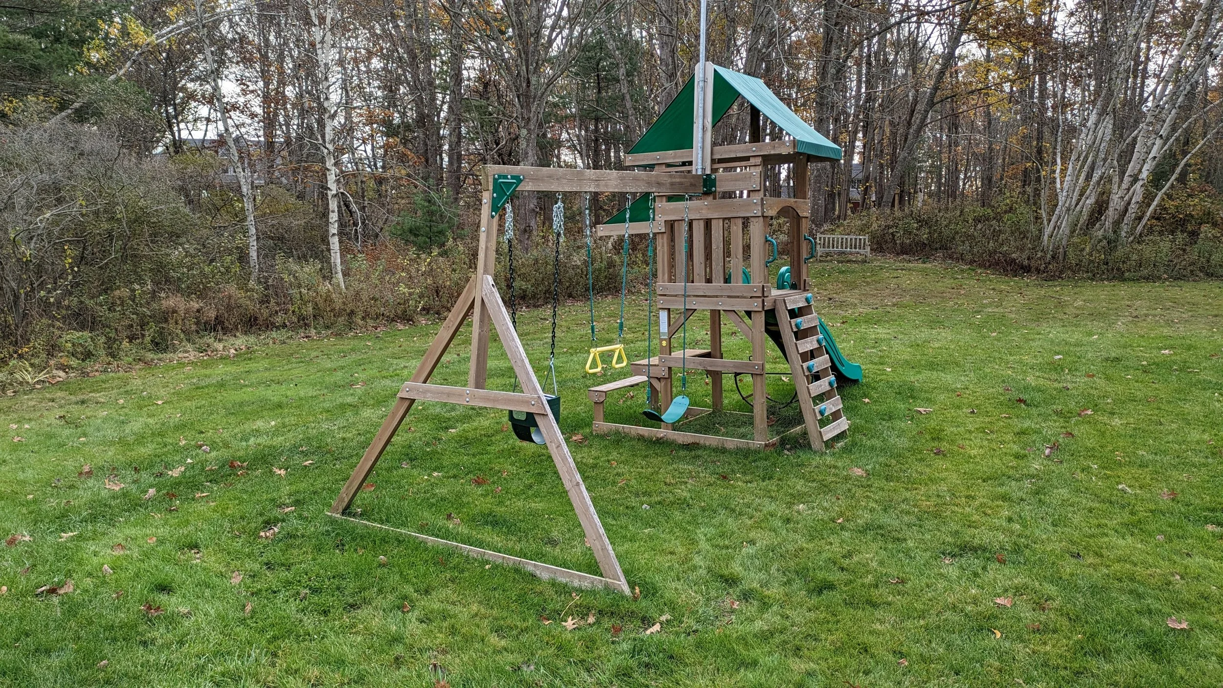 Wooden playground set with swings, slide, climbing wall, and a small covered platform on a grassy lawn with trees in the background.
