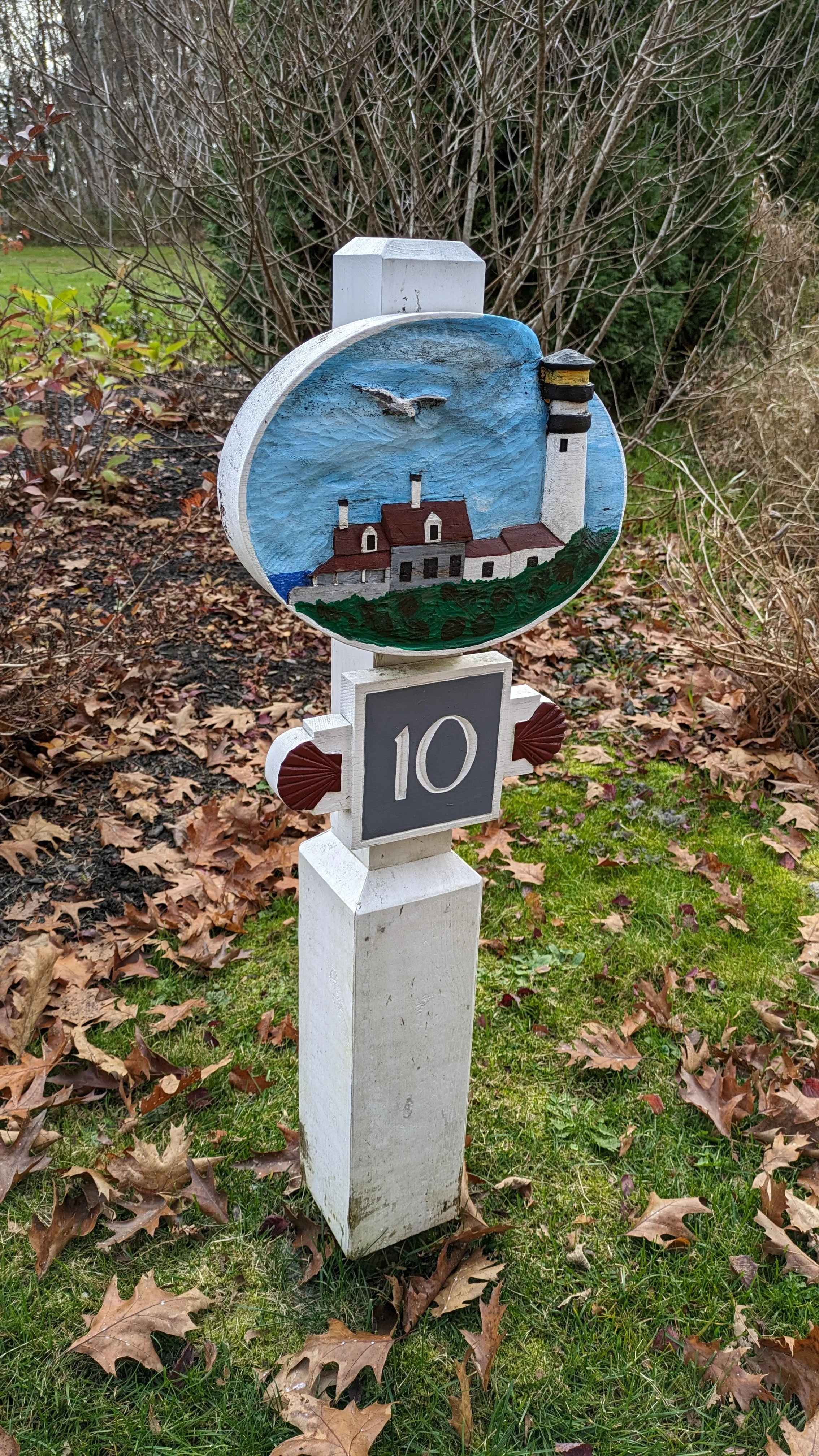 Decorative white wooden post with a painted lighthouse scene and the number 10, situated in a garden with fallen leaves and bushes in the background.