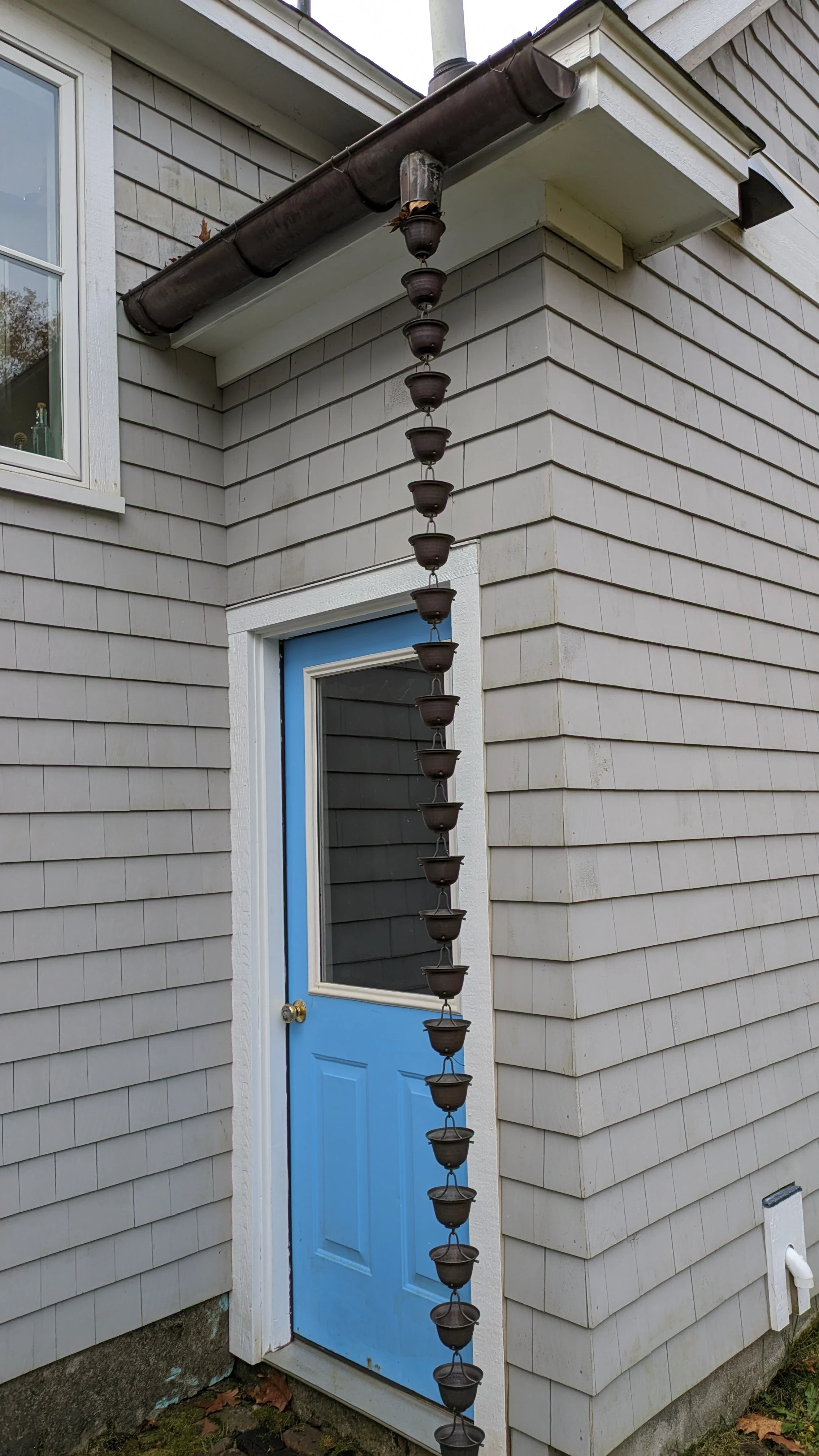 Downspout with water cups running from the gutter to the door, resembling a water cascade outside a house with gray shingles and a blue door.