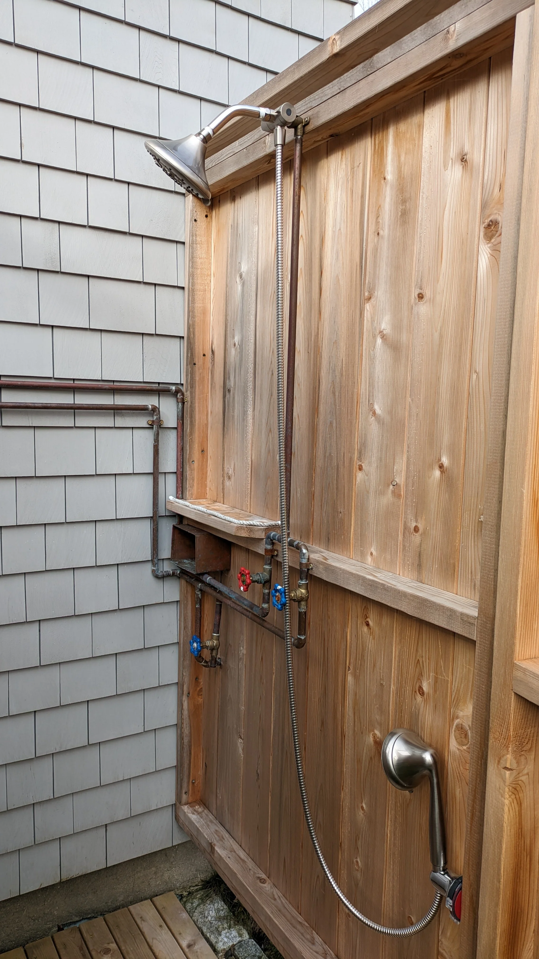 An outdoor shower setup with a metal showerhead, a flexible hose, and two water valves mounted on a wooden wall, next to a house with gray siding.