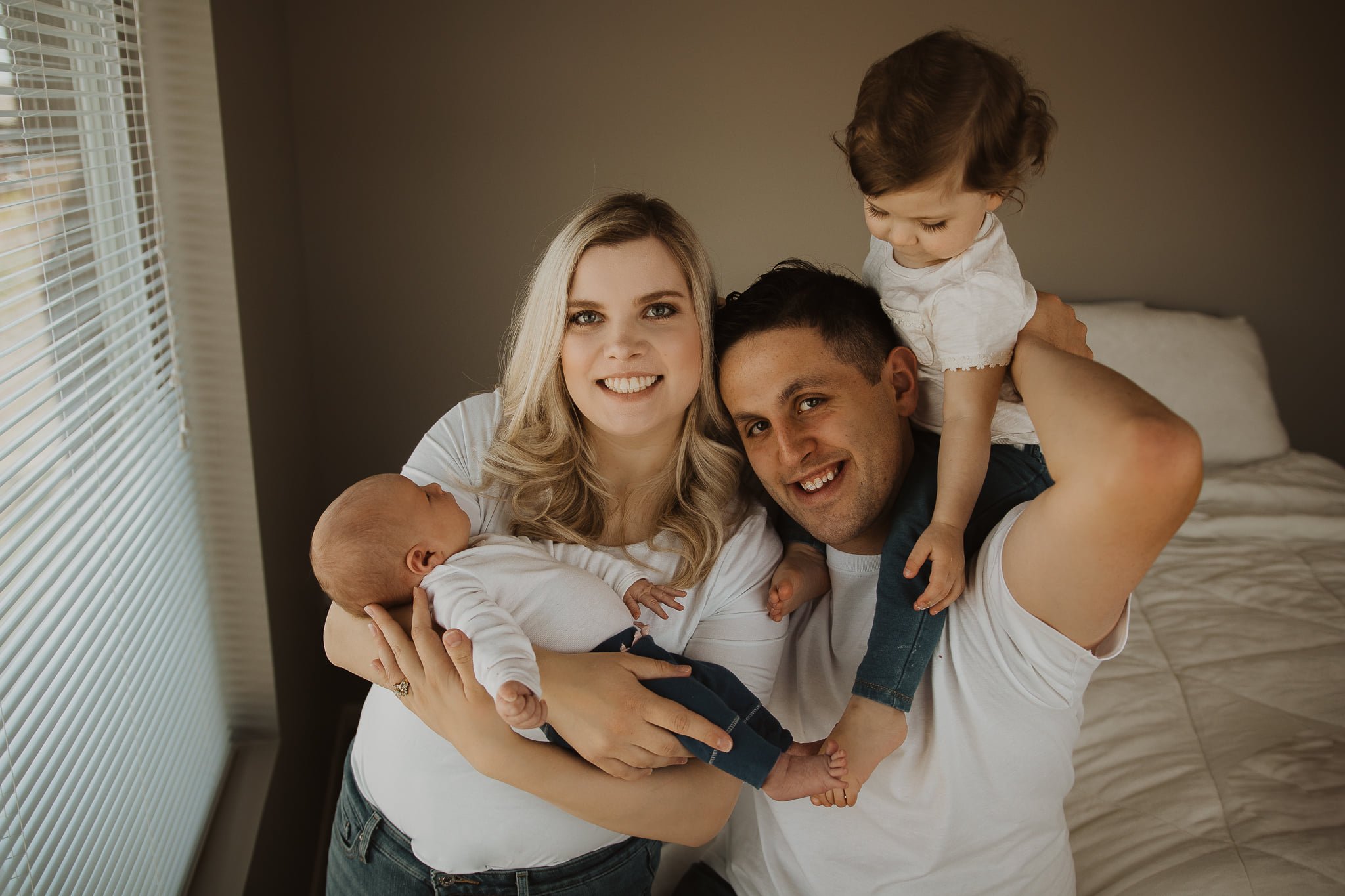 A family of four smiling and holding their children, a newborn and a toddler, in a bedroom with beige walls and a window with blinds.