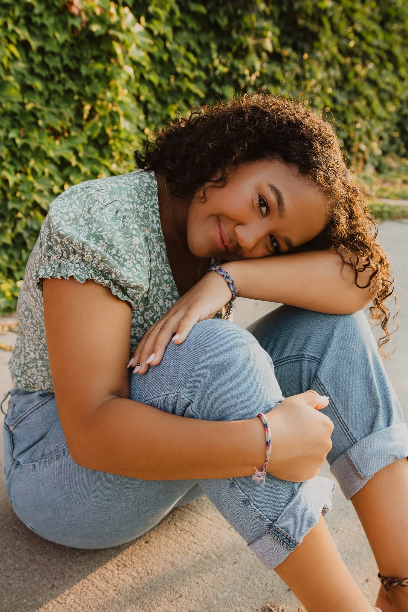 A young woman with curly hair, wearing a green floral blouse and light jeans, sitting on the ground with her knees up, smiling at the camera in front of a leafy green hedge.