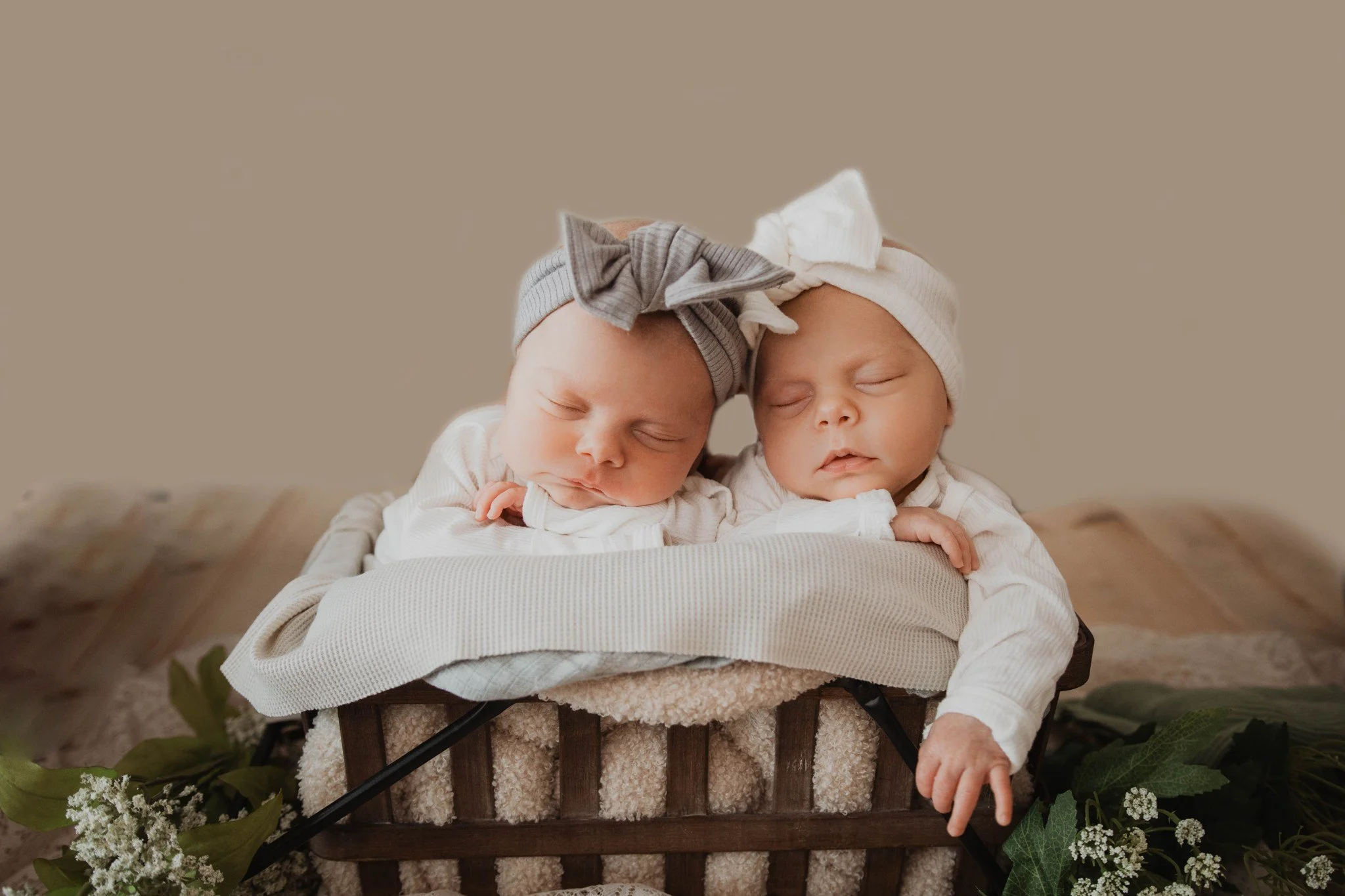 Two sleeping babies in a wicker basket with beige blankets, wearing matching white outfits and headbands with bows, surrounded by green leaves and small white flowers.