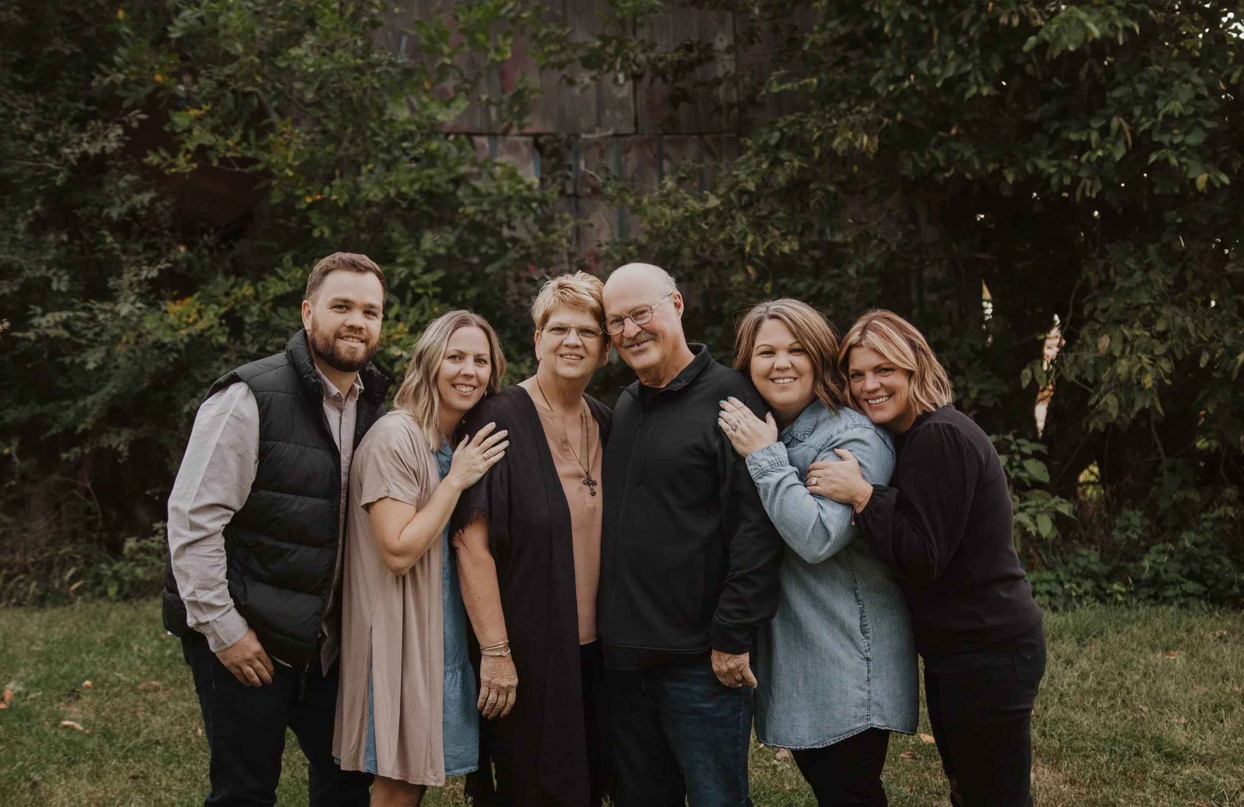 Group of six people standing outdoors in front of greenery and a wooden structure, smiling and hugging each other.