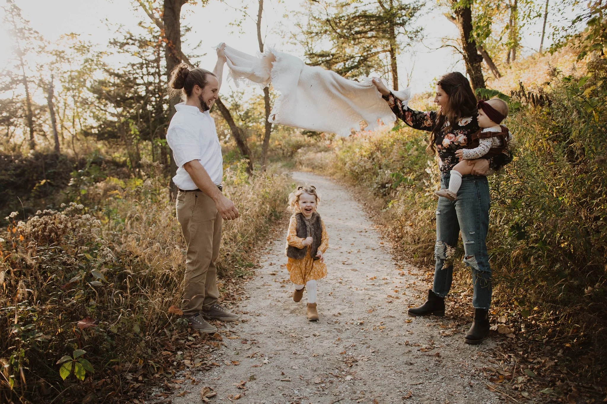 A family of four enjoying a walk in a wooded area during autumn, with a man, a woman holding a toddler, and a young girl running happily on a dirt path.