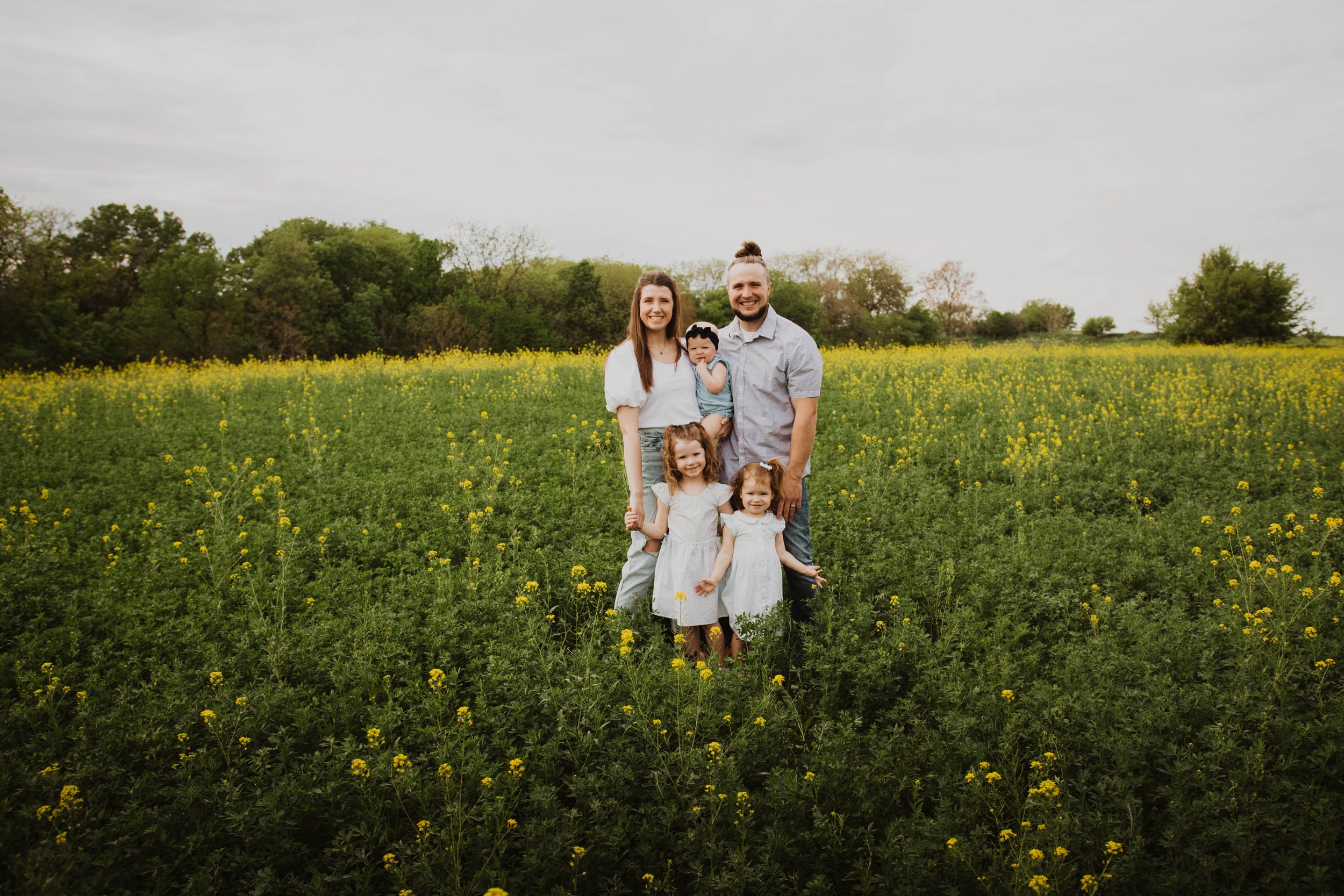 A family of six standing in a yellow flower field with green trees in the background, smiling at the camera.