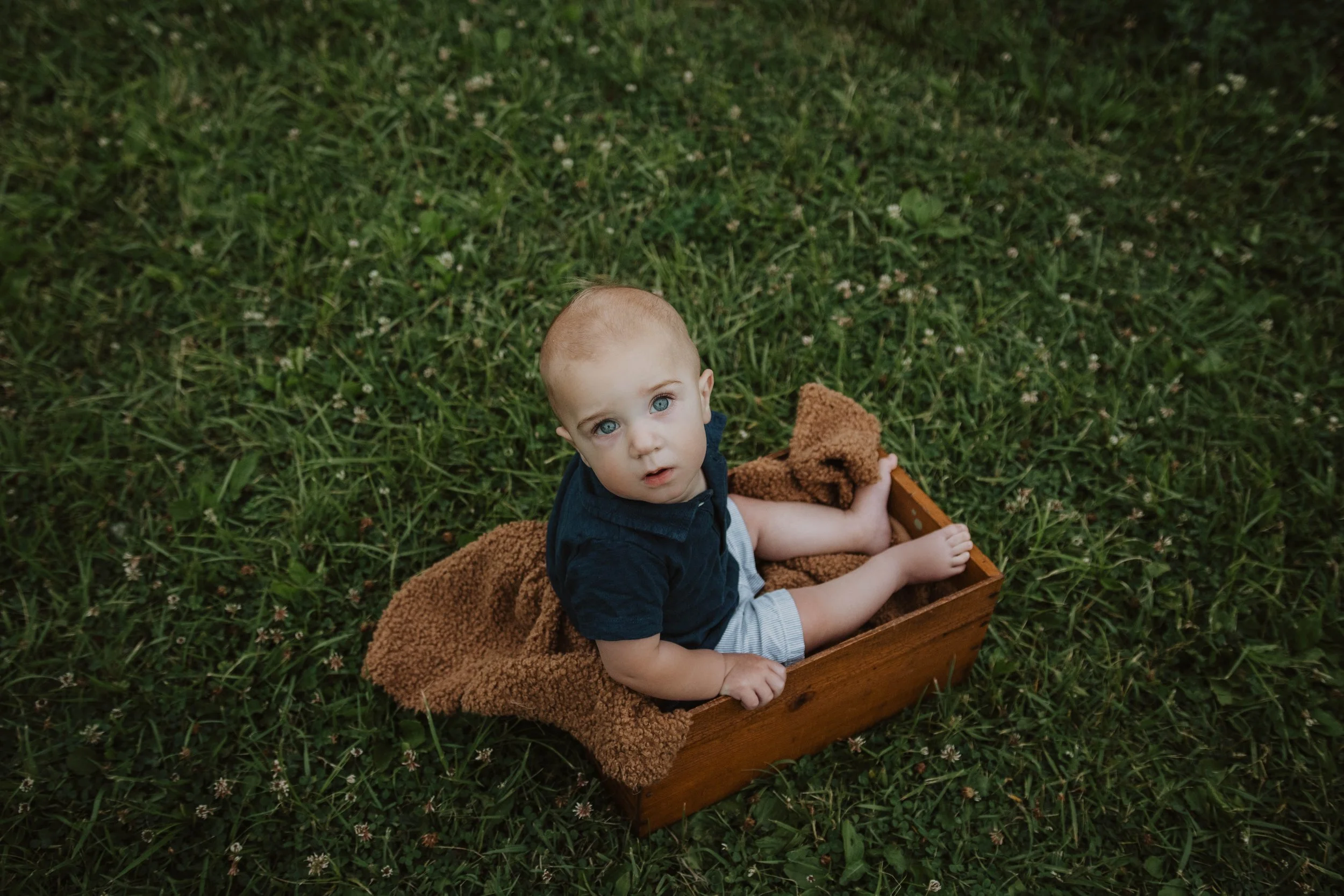 A young child with blue eyes and a shaved head sitting in a wooden box on grass, looking up at the camera with a curious expression.