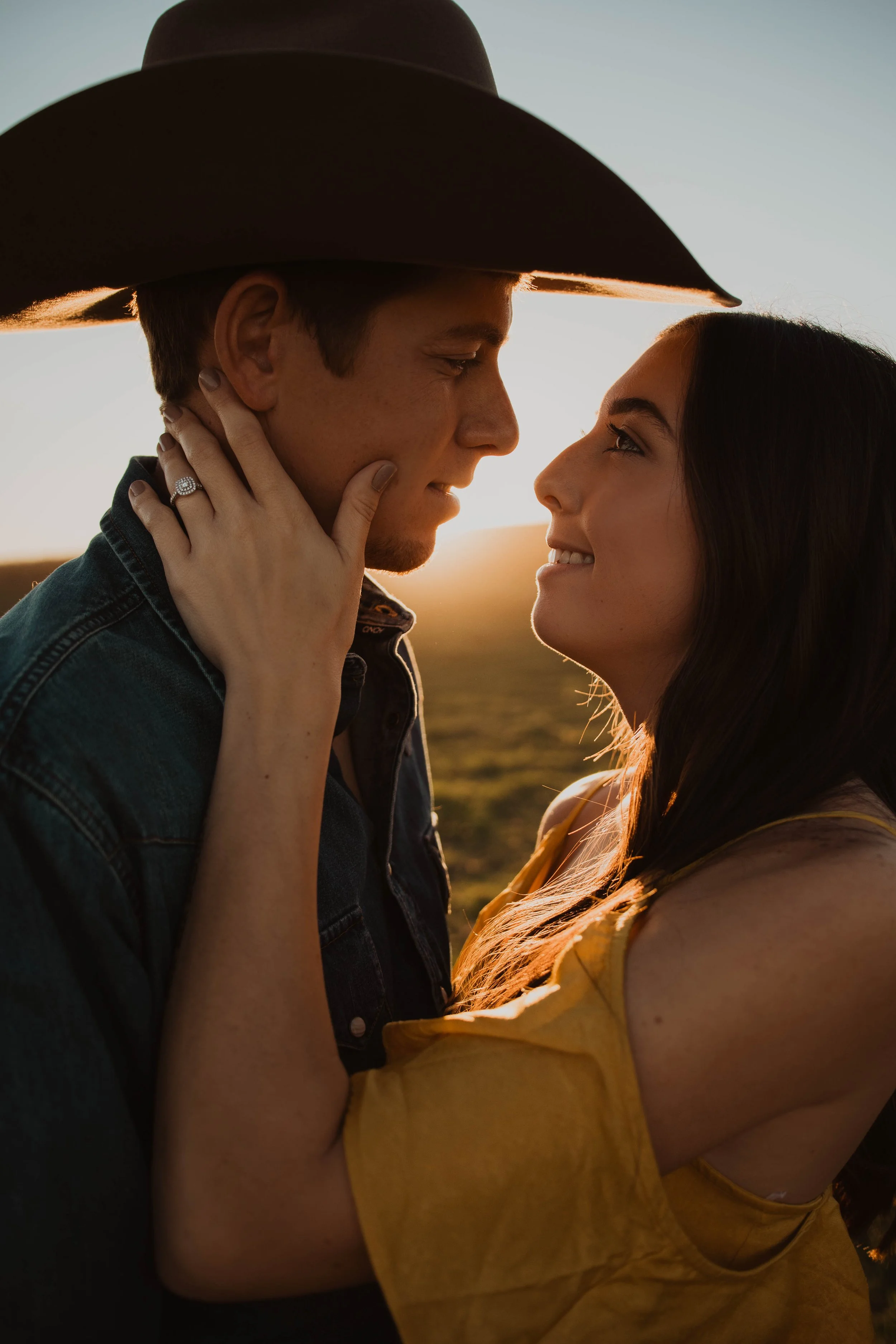 A young man and woman share an intimate moment outdoors at sunset, with their faces close together and a warm glow on their skin. The woman is wearing a yellow top and has dark hair, while the man is wearing a denim jacket and a large dark hat. The w