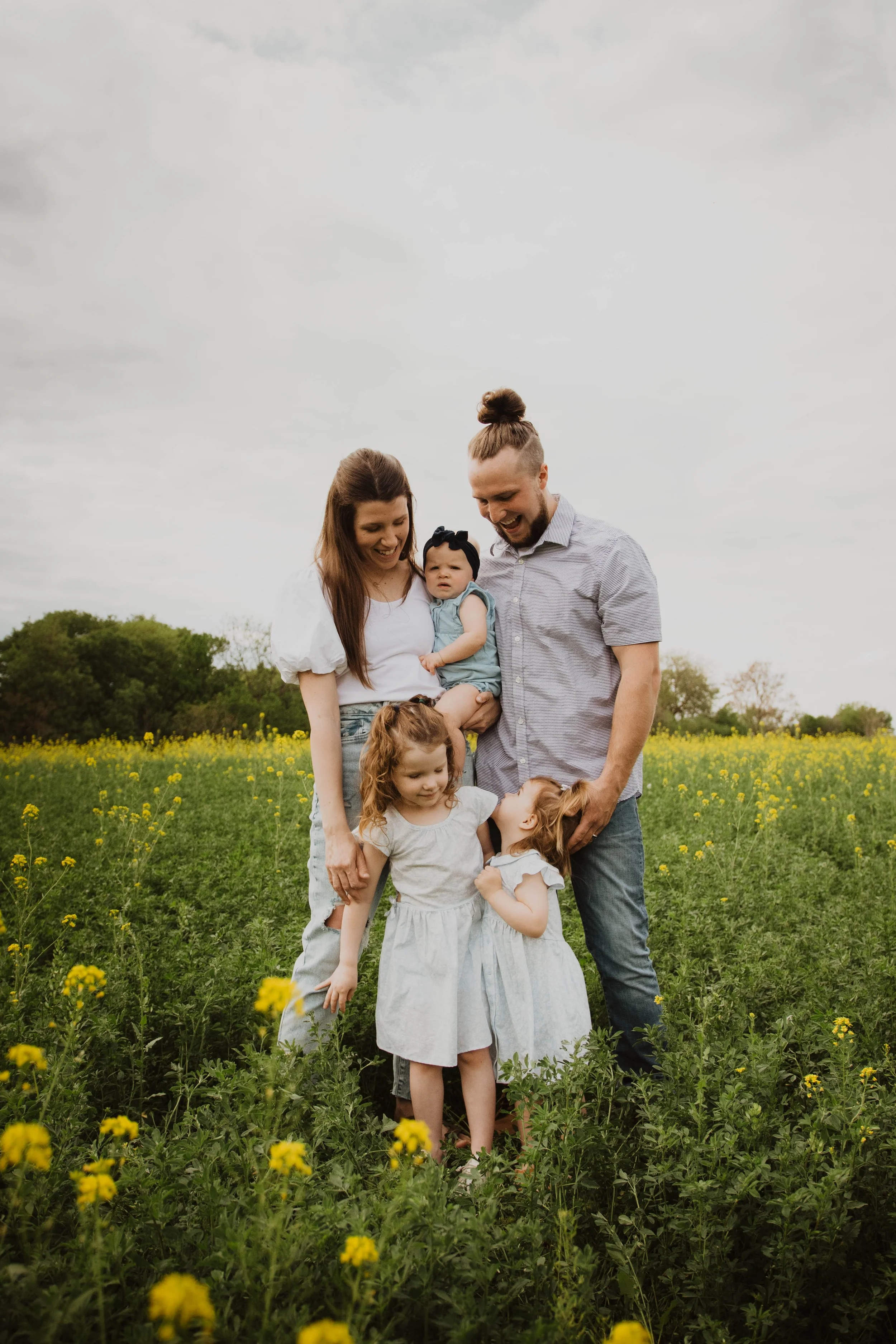 A family of five, including a mother, father, and three young daughters, standing together in a yellow flower field on a cloudy day, smiling and interacting.