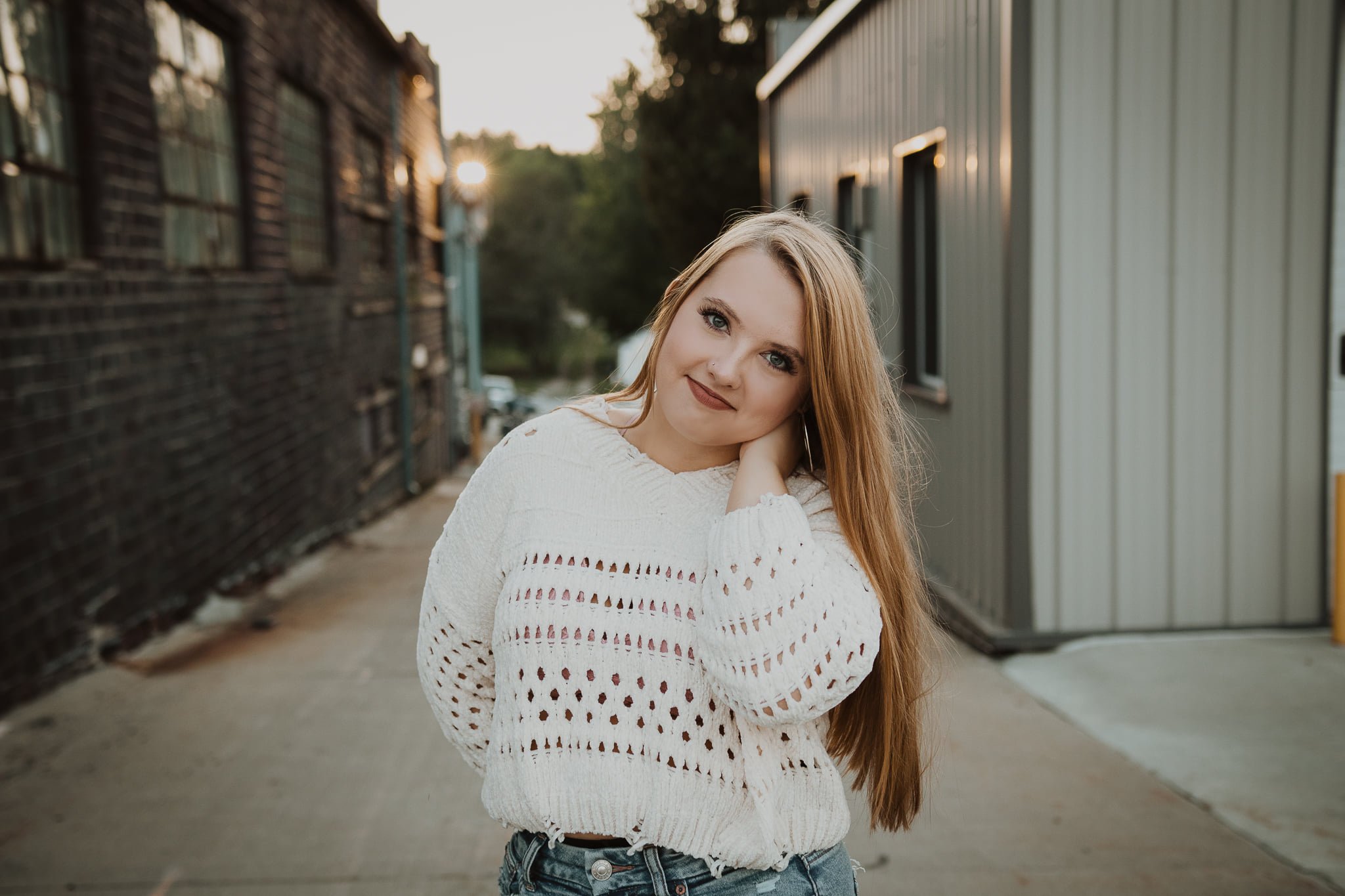 A young woman with long red hair and blue eyes posing outdoors in front of buildings during sunset, wearing a white crochet sweater and jeans.