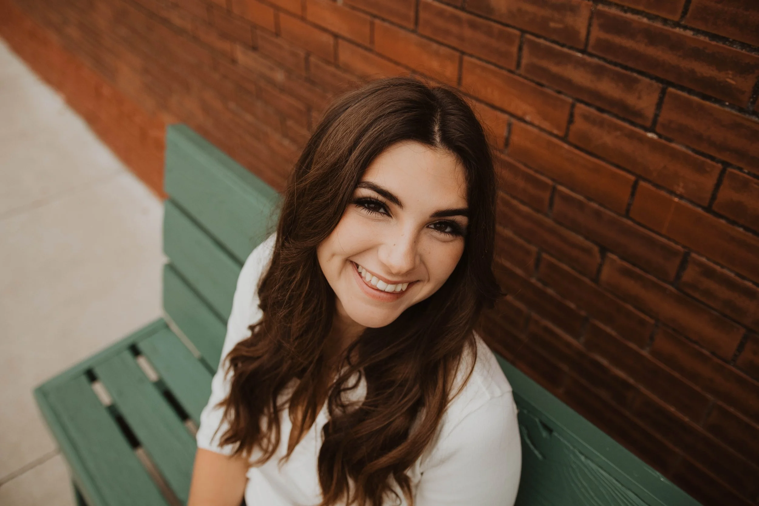 A smiling young woman with long brown hair sitting on a green bench against a red brick wall.