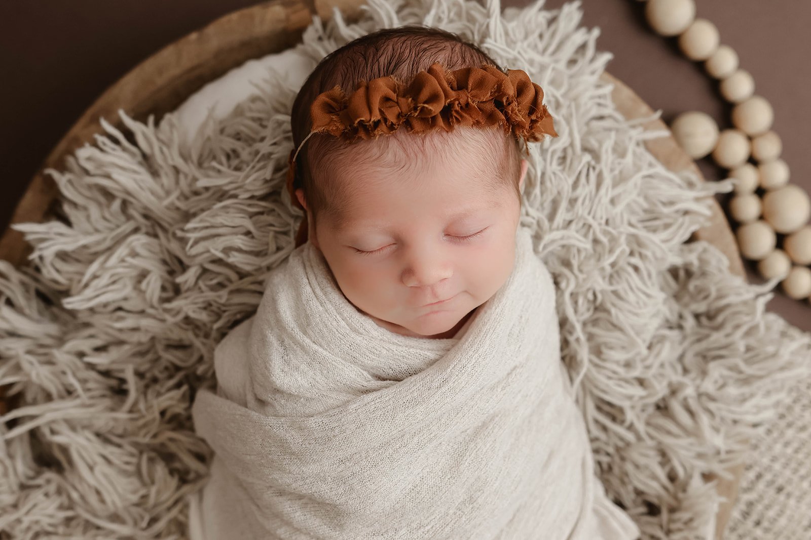 A sleeping newborn baby wrapped in a beige blanket, wearing a brown ruffled headband, lying on a fluffy cream-colored blanket on a round wooden surface.