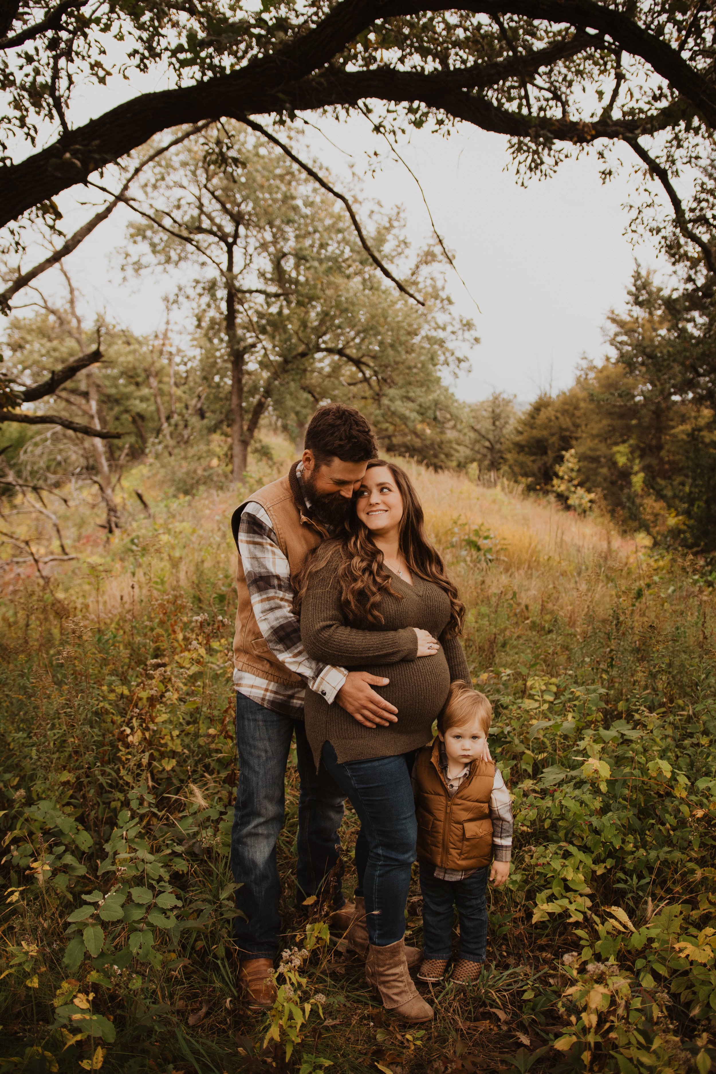 A family of three standing in a forested area during autumn; a pregnant woman with long brown hair, a man with a beard, and a young boy with light hair wearing autumn clothing.