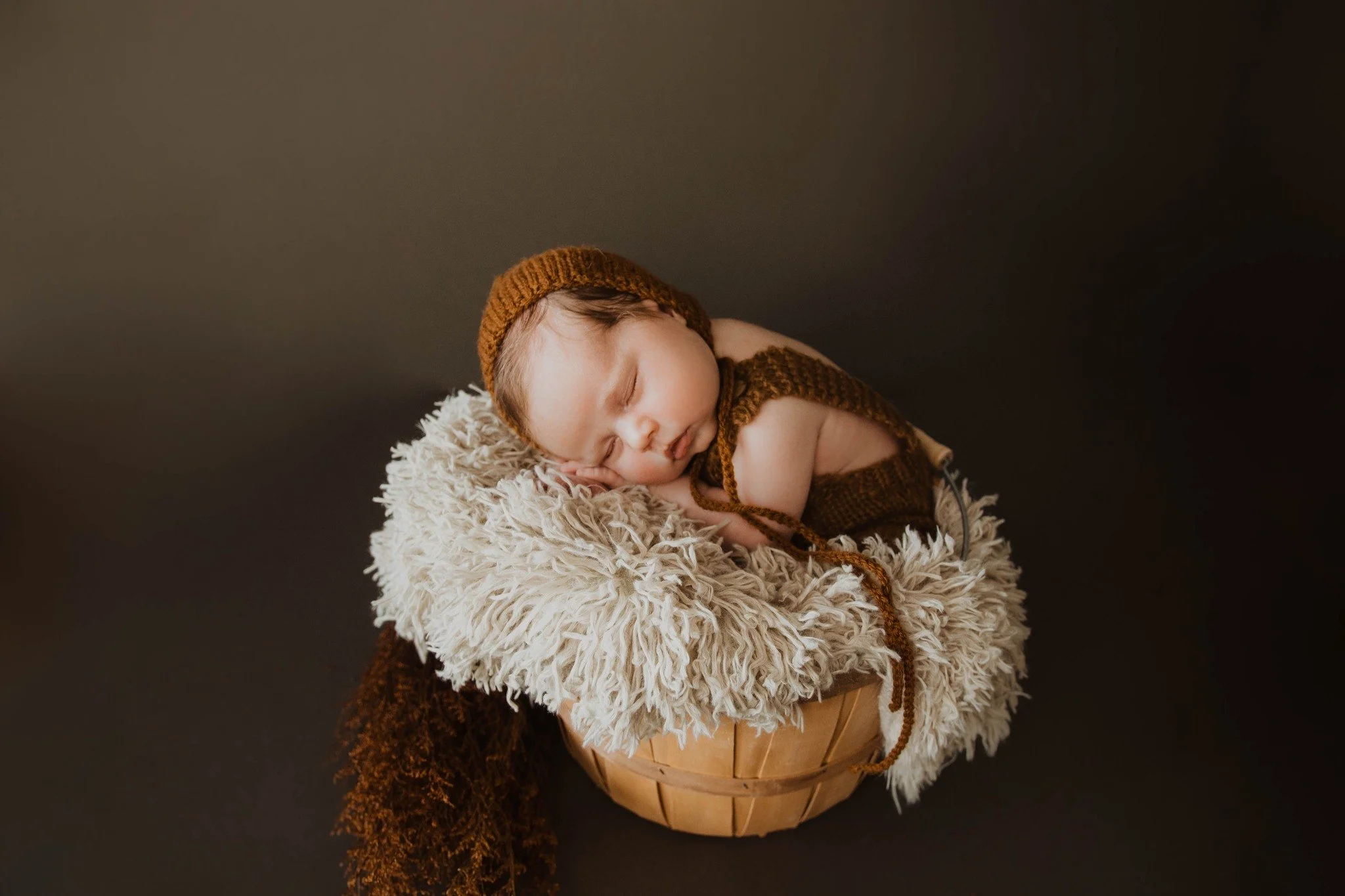 A sleeping baby in a brown knitted hat, nestled in a soft, fluffy blanket inside a woven basket against a dark background.
