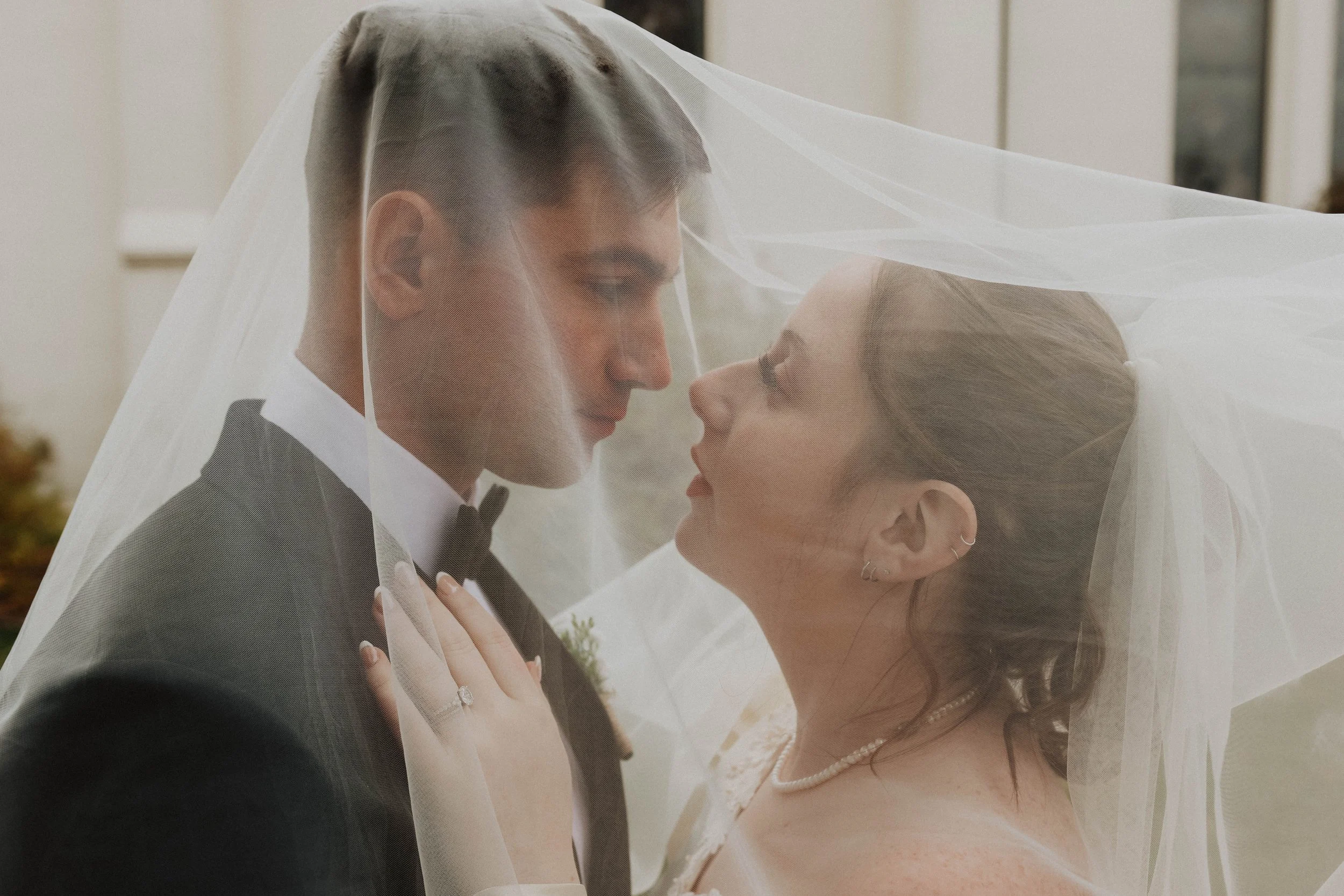 A bride and groom under a wedding veil, gazing at each other, with the groom in a tuxedo and the bride in a wedding dress, outside a house.