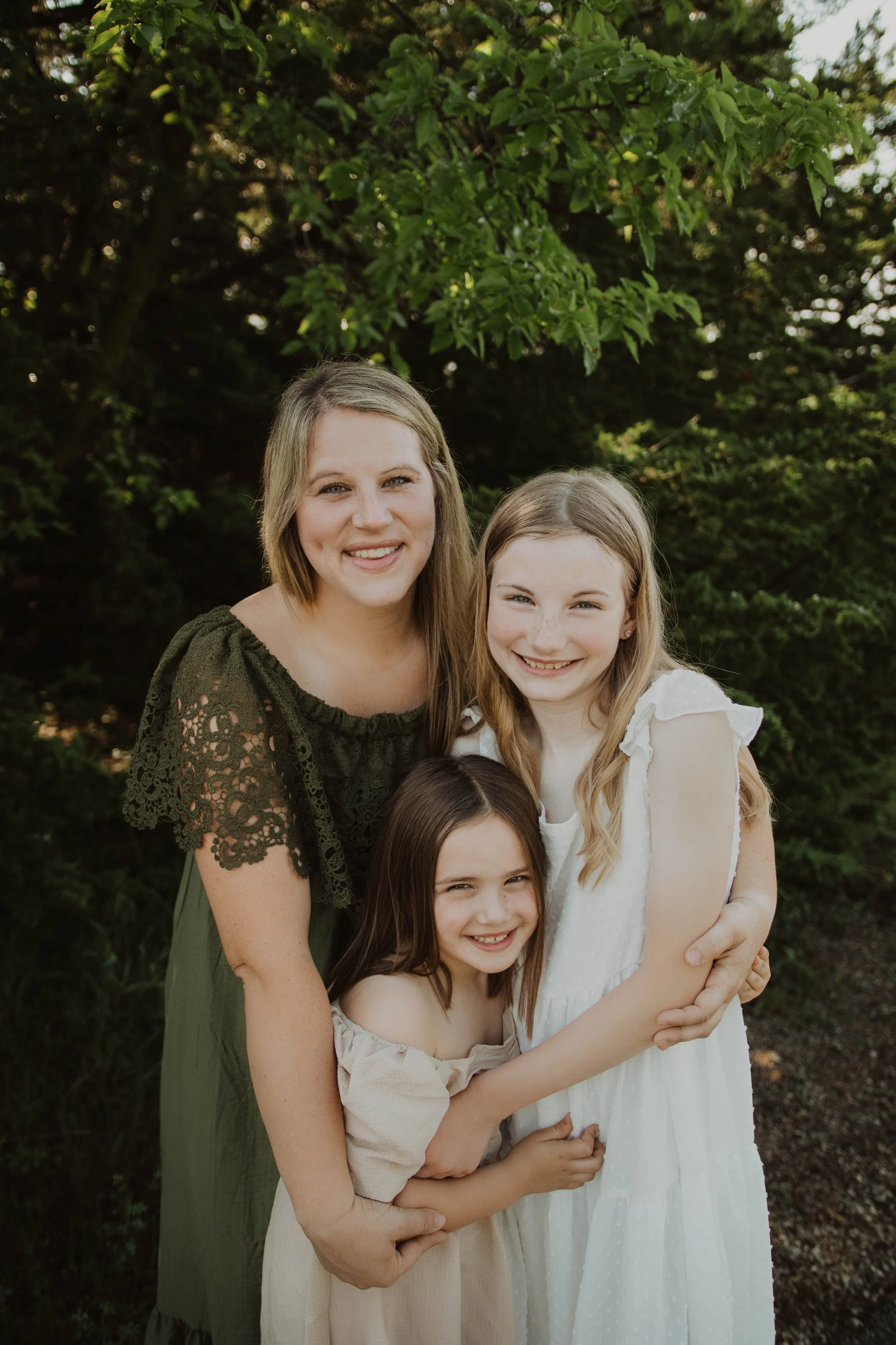 A woman and two young girls smiling and hugging outdoors, surrounded by green foliage.