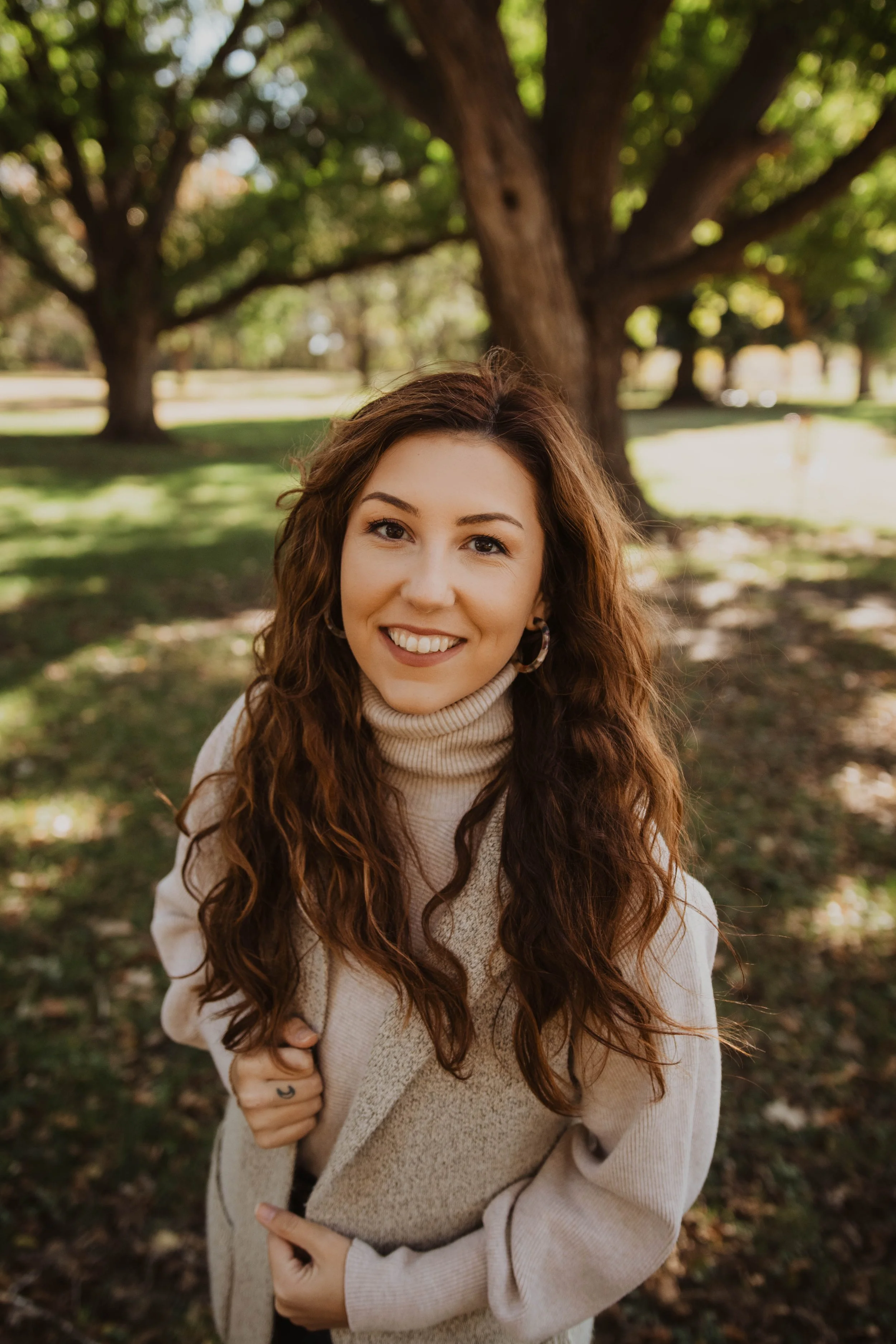 A woman with long, wavy brown hair smiling outdoors in a park with trees and green grass.