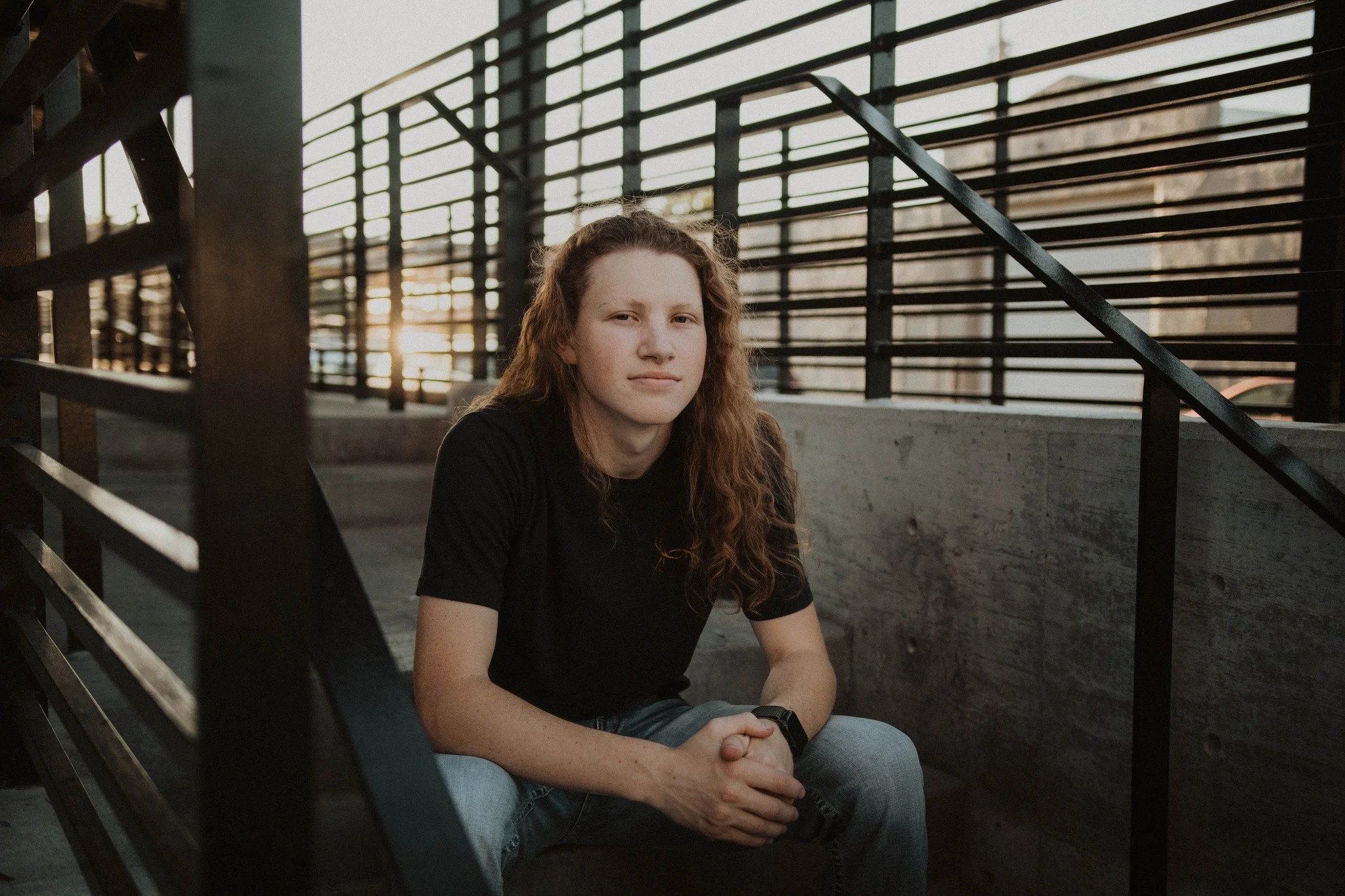 A young woman with curly red hair sitting on concrete stairs outdoors, looking into the camera with a neutral expression, during sunset.