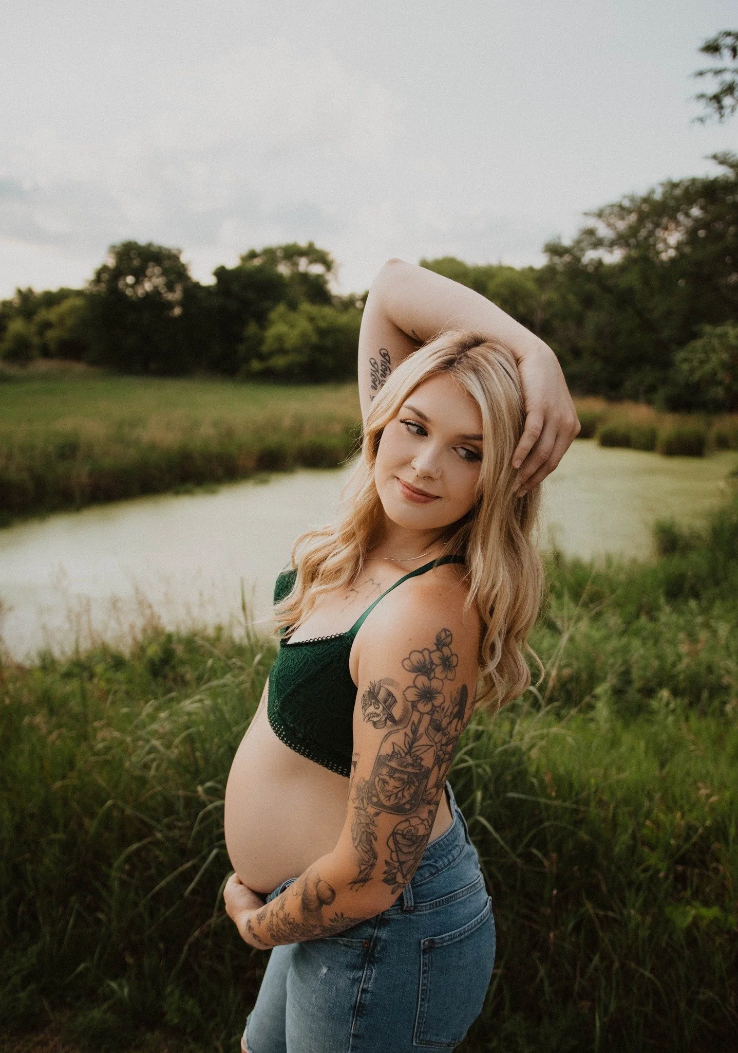A pregnant woman with long blonde hair, tattoos on her arm, wearing a black and green top and denim shorts, standing outdoors near a pond with trees in the background.