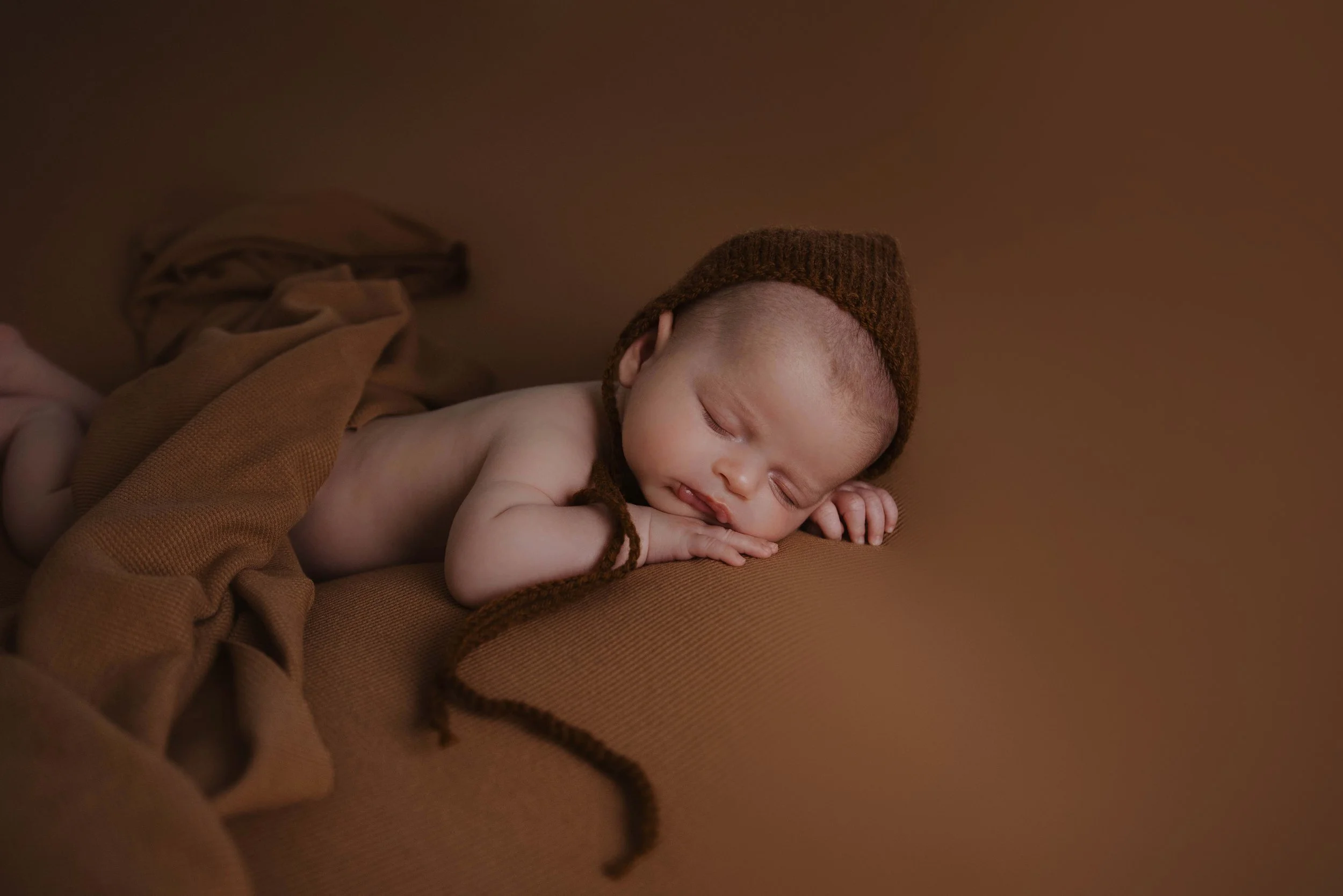 A sleeping baby lying on a brown surface, wearing a brown knit hat and brown blanket, with arms folded under the head.