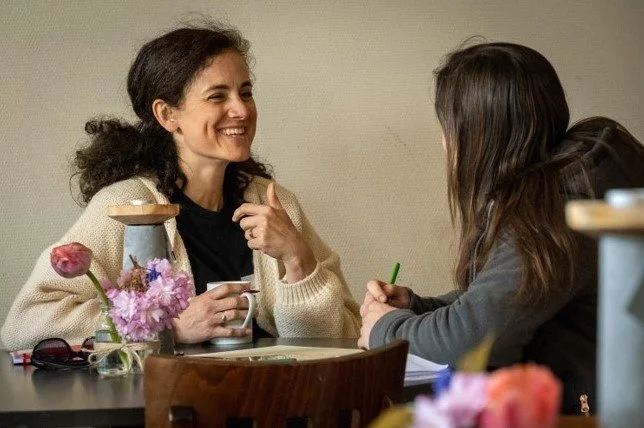 Twee vrouwen hebben een gesprek aan een tafel met bloemen en een kopje koffie.