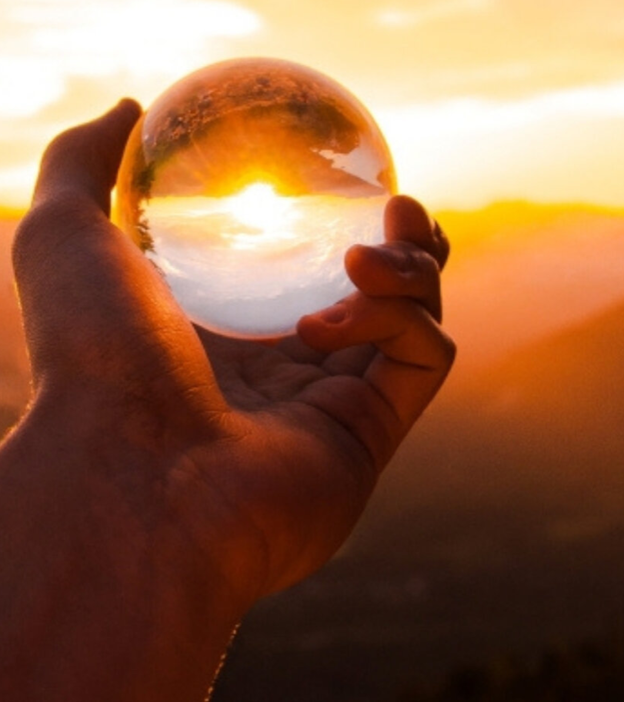 Hand die een glazen bol vasthoudt met een zonsondergang en de lucht erin weerspiegelend.